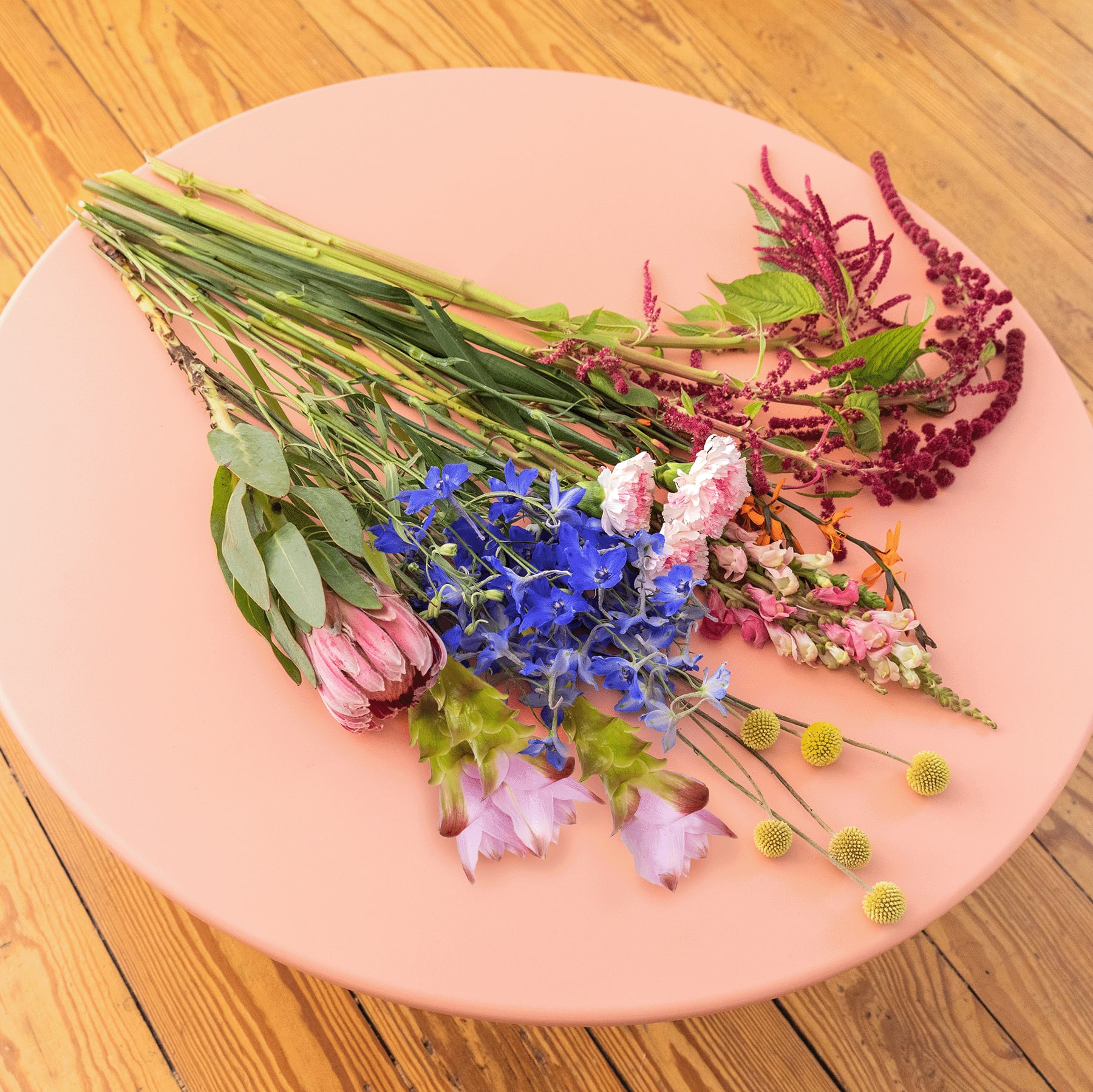 Ein lebendiges Arrangement aus verschiedenen bunten Blumen in Blau-, Rosa- und Grüntönen liegt auf einem rosafarbenen runden Teller vor einem hölzernen Hintergrund.