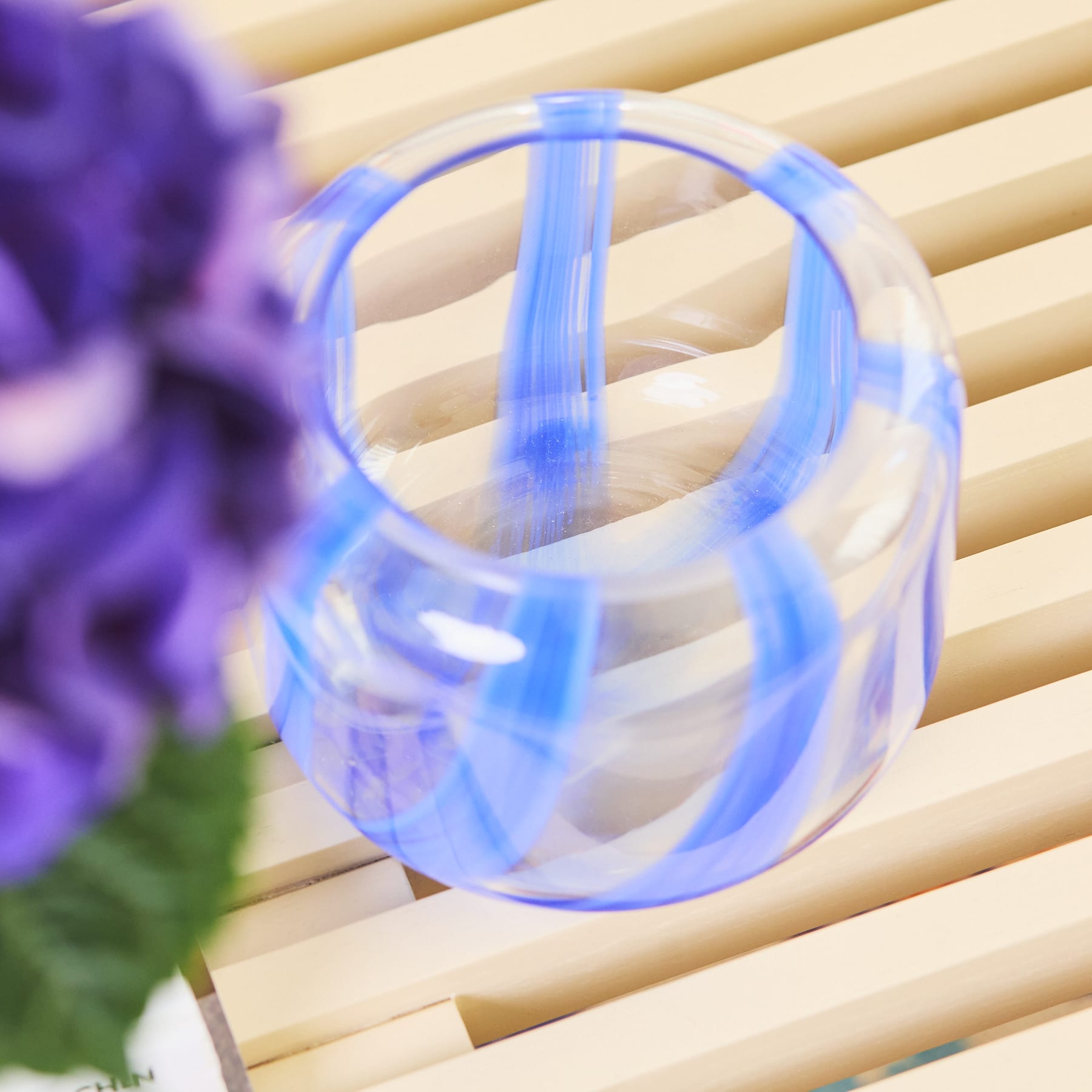 Eine durchsichtige blaue Glasvase oder ein Behälter steht auf einer Holzoberfläche, im Hintergrund sind violette Blumen zu sehen.
