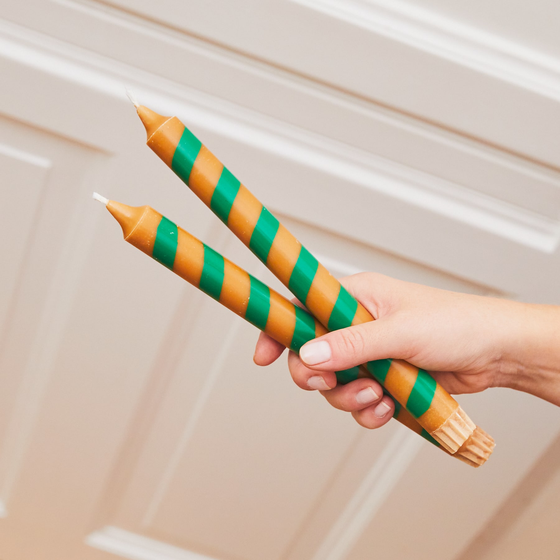 Das Bild zeigt eine Hand, die ein Paar gestreifte Eistüten hält, vor einem weißen Hintergrund und etwas, das wie eine Holzoberfläche oder ein Türrahmen aussieht.