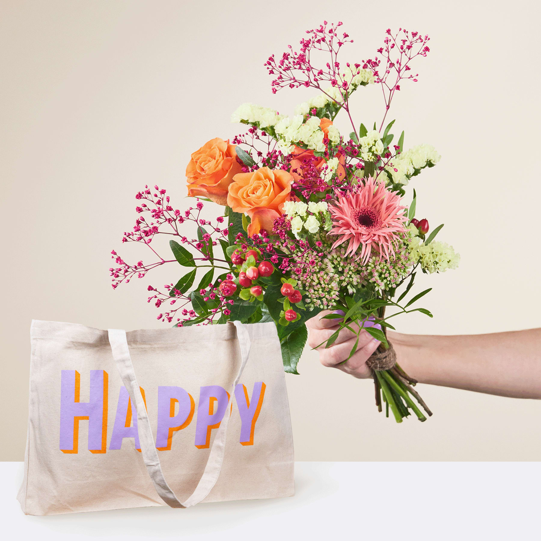 Eine Person hält einen leuchtend bunten Blumenstrauß, darunter orangefarbene Rosen, rosa Gerbera und zartes Schleierkraut, in den Händen, zusammen mit einer wiederverwendbaren Einkaufstasche mit dem Aufdruck „Happy“ vor einem schlichten Hintergrund.