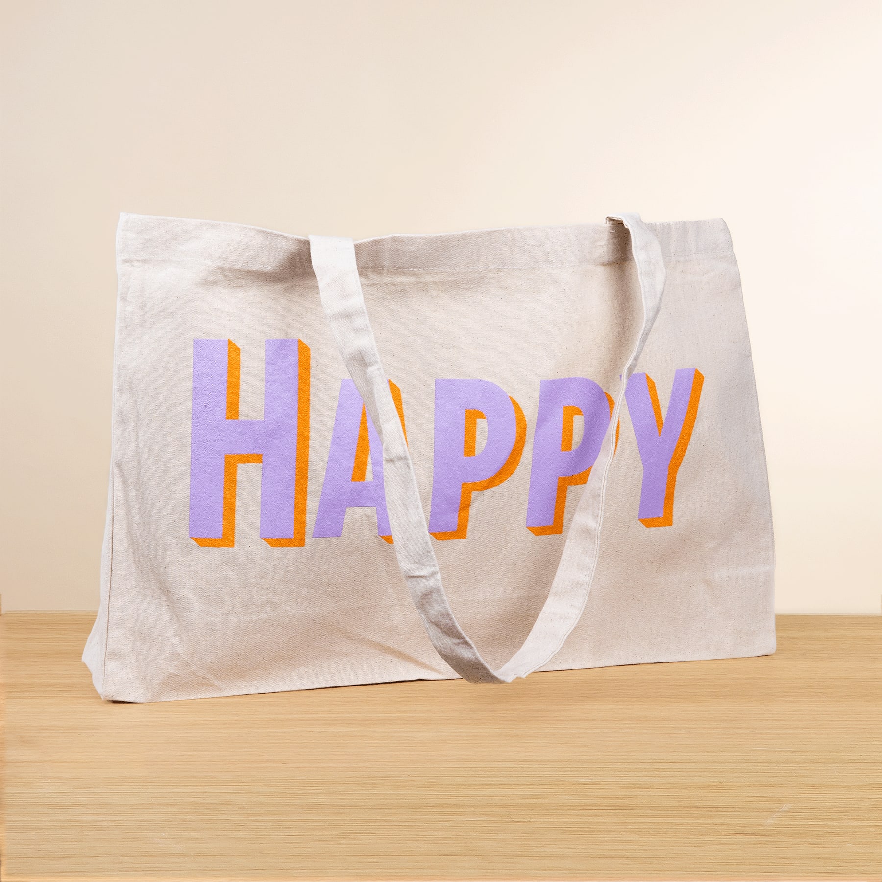 Eine weiße Einkaufstasche aus Segeltuch mit dem Wort „HAPPY“ in bunten Buchstaben liegt auf einer Holzoberfläche vor einem schlichten Hintergrund.