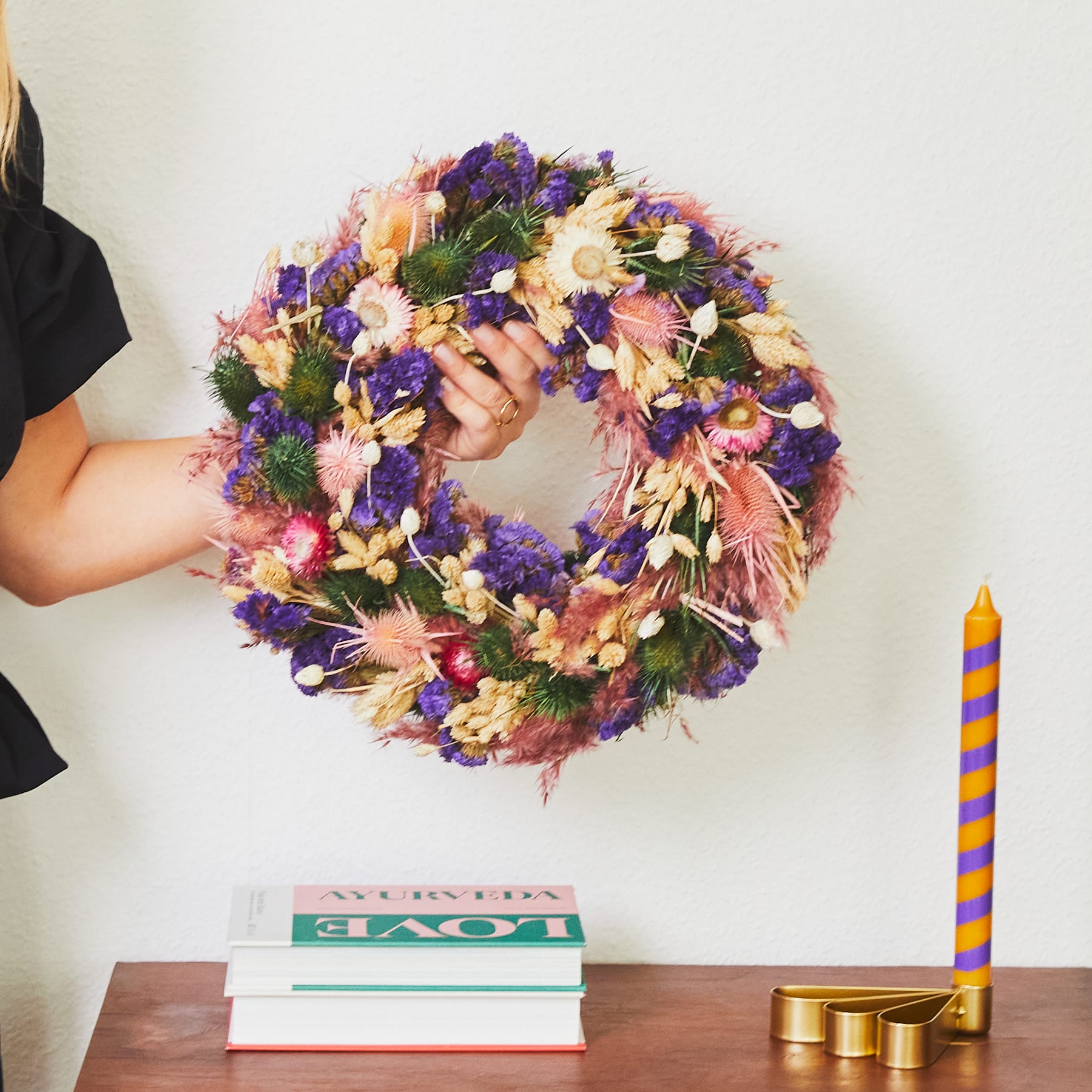 Im Vordergrund steht ein farbenfroher Blumenkranz aus Blumen in verschiedenen Lila- und Rosatönen, der auf einem Stapel Bücher ruht und von einer gestreiften Kerze begleitet wird.