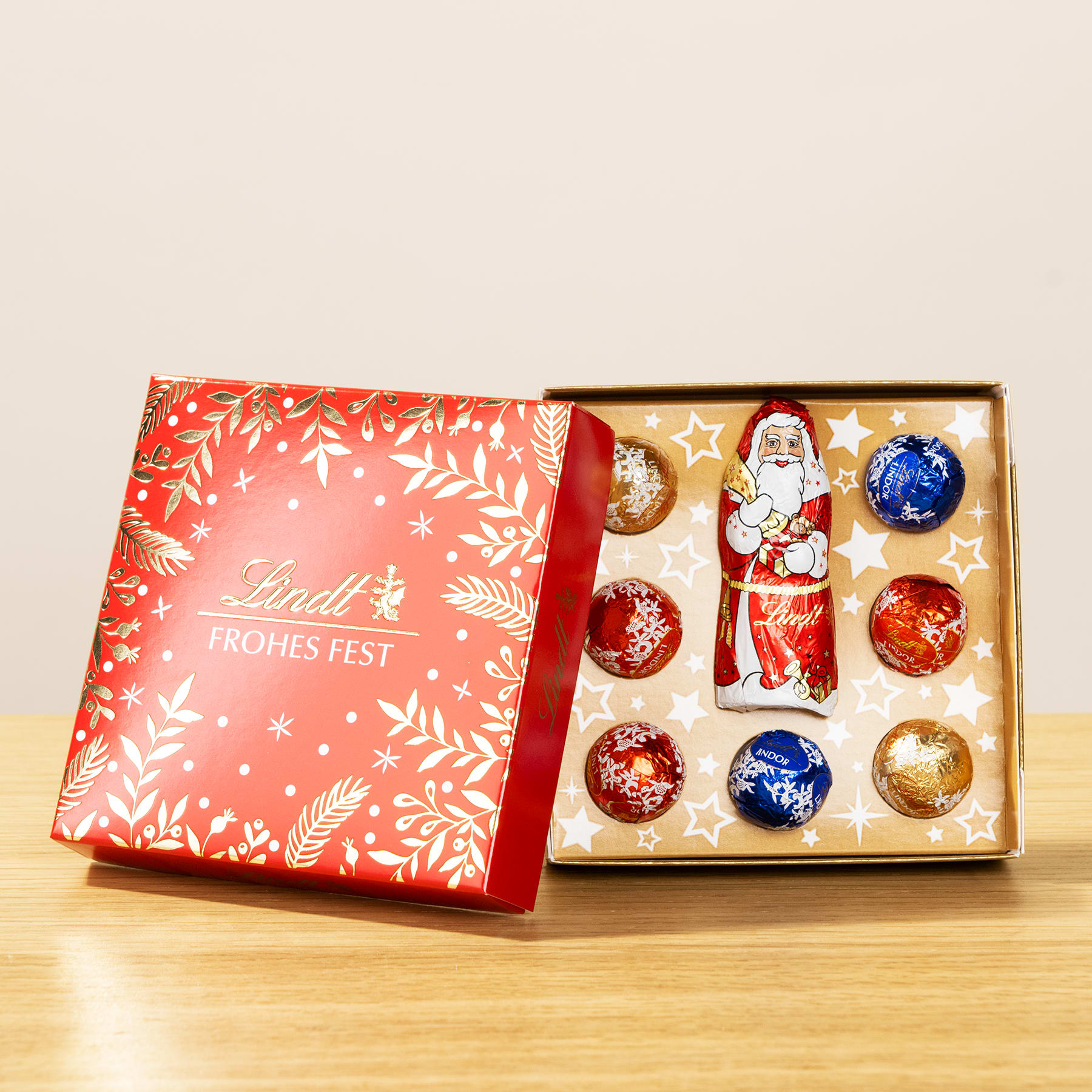 Auf einer Holzoberfläche steht eine rot-weiße Geschenkbox mit verschiedenen weihnachtlichen Schokoladenbonbons und Dekorationen.
