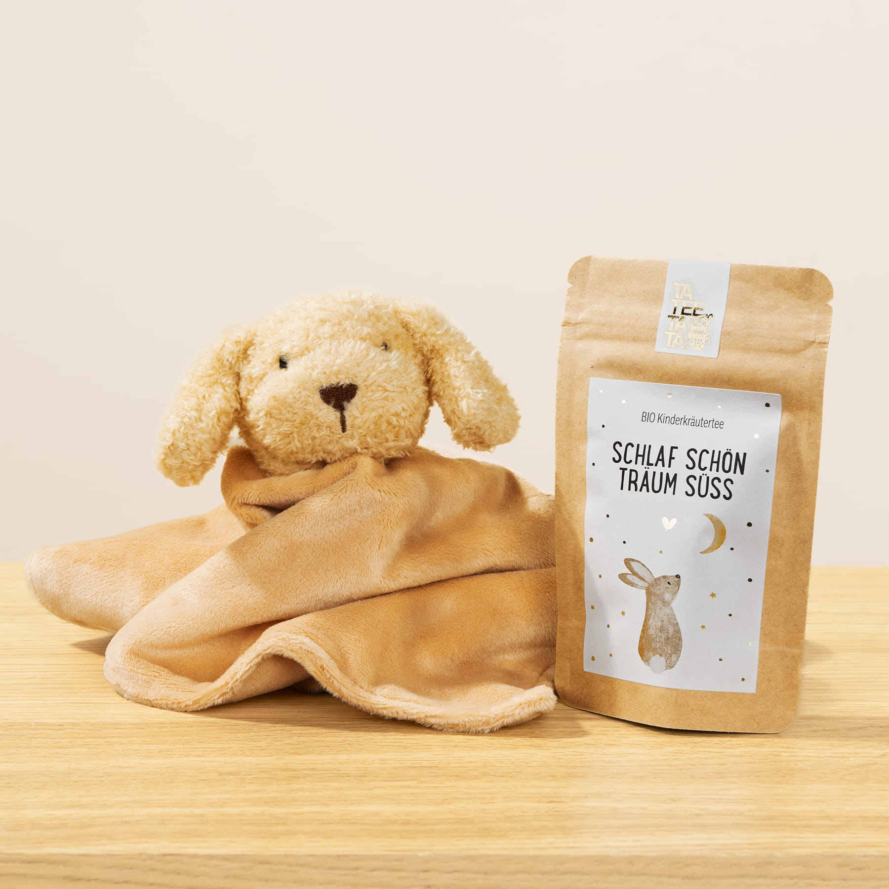 Auf einer Holzfläche sitzt ein weicher Plüschhund oder ein Stoffkaninchen neben einer Papiertüte mit der Aufschrift „Schlaf schon traum süss“.
