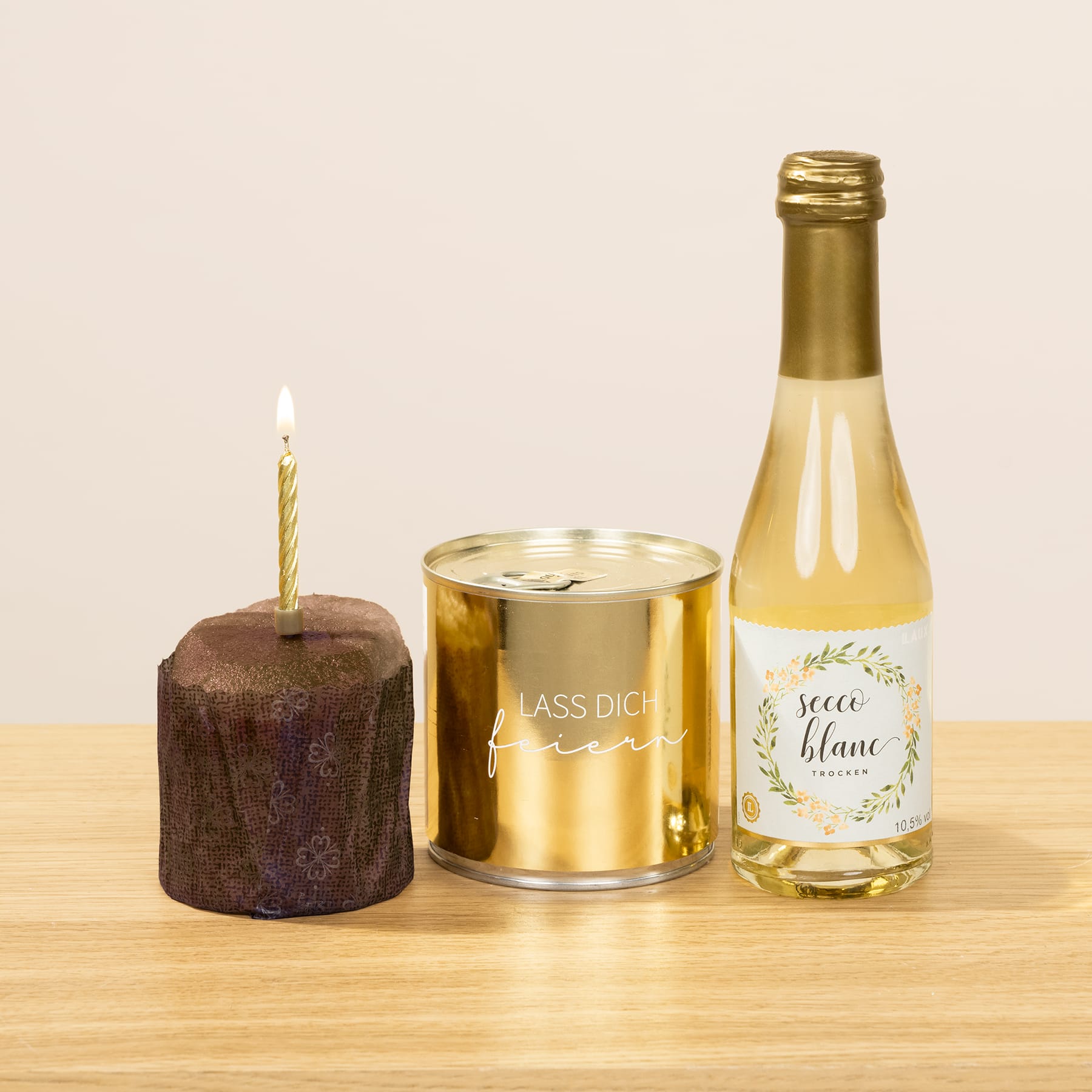 Auf einem Holztisch stehen eine Geburtstagstorte mit einer brennenden Kerze, eine goldfarbene Dose und eine Flasche Weißwein.
