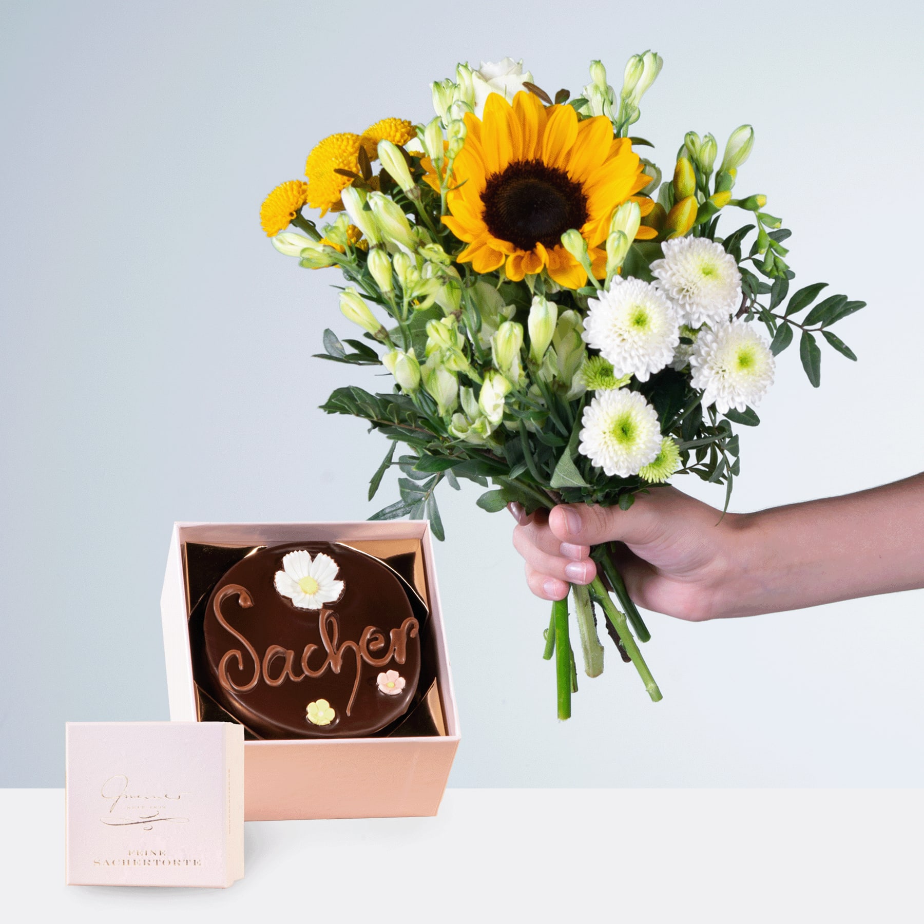 Eine Person hält einen Strauß leuchtender Sonnenblumen, Gänseblümchen und anderer Blumen in den Händen, neben einer Schokoladengeschenkschachtel mit der Aufschrift „Sachet“.