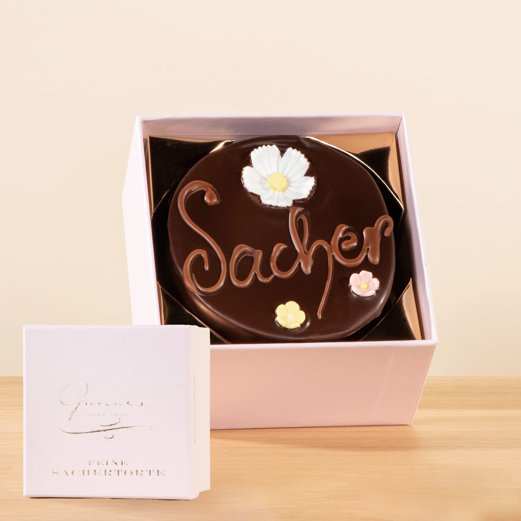 Eine Schokoladentorte mit der Aufschrift „Sacher“, dekoriert mit weißen Blumen, steht in einer rosa Schachtel auf einer Holzoberfläche.