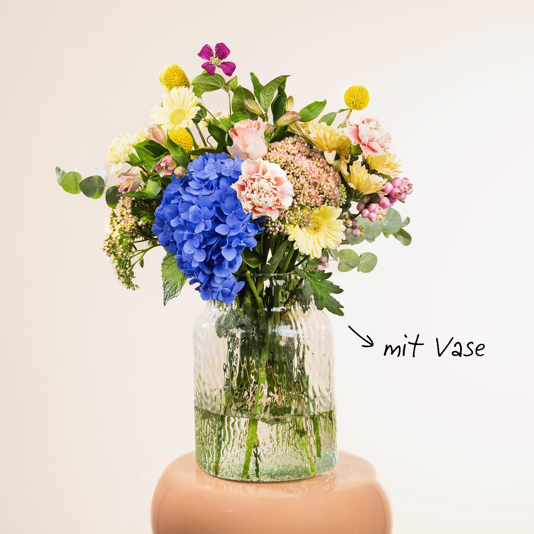Ein leuchtender Strauß bunter Blumen, darunter blaue Hortensien, gelbe Narzissen und rosa Nelken, wird in einer Glasvase auf einer hellen Oberfläche präsentiert.