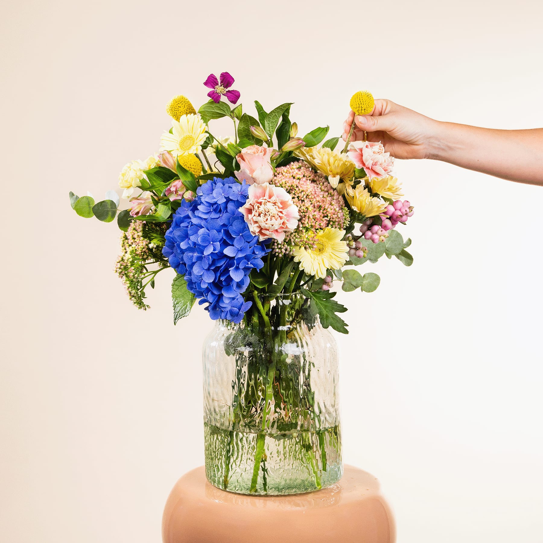 Ein leuchtender Strauß bunter Blumen, darunter blaue Hortensien, gelbe Narzissen und rosa Nelken, steht in einer Glasvase vor einem schlichten, hellen Hintergrund.