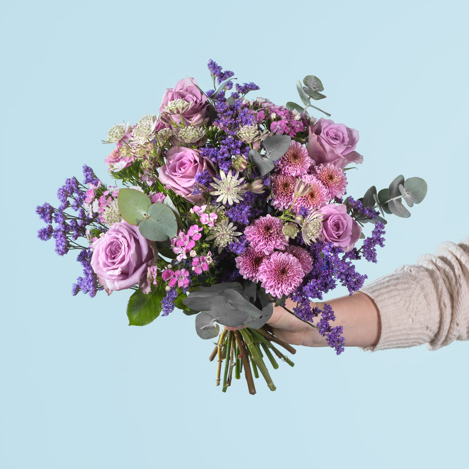 Ein leuchtender Strauß aus rosa Rosen, violetten Blumen und Grünpflanzen, den eine Person vor einem hellblauen Hintergrund in den Händen hält.