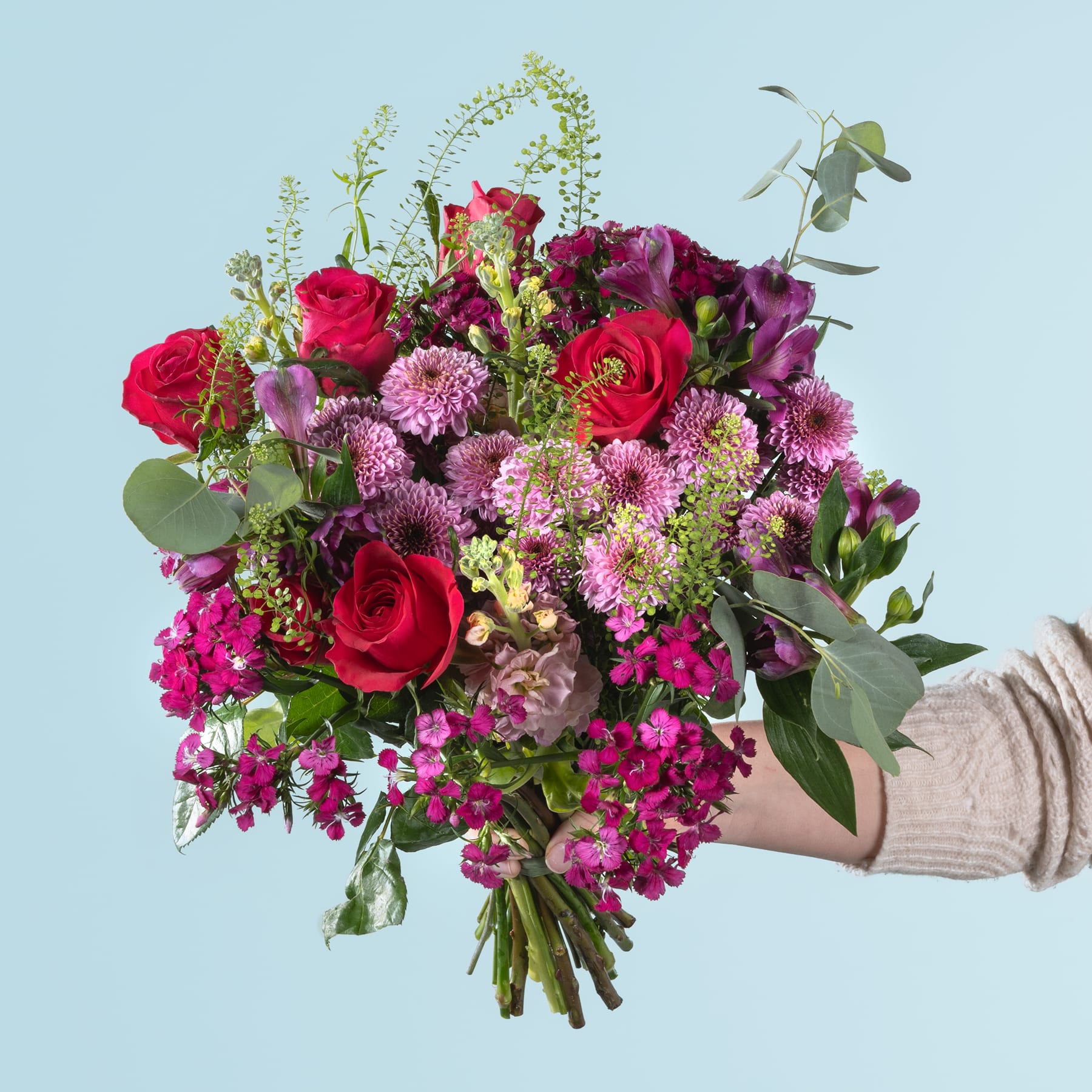 Ein leuchtender Strauß roter Rosen, rosa Chrysanthemen und anderer bunter Blumen, den eine Person vor einem hellblauen Hintergrund in den Händen hält.