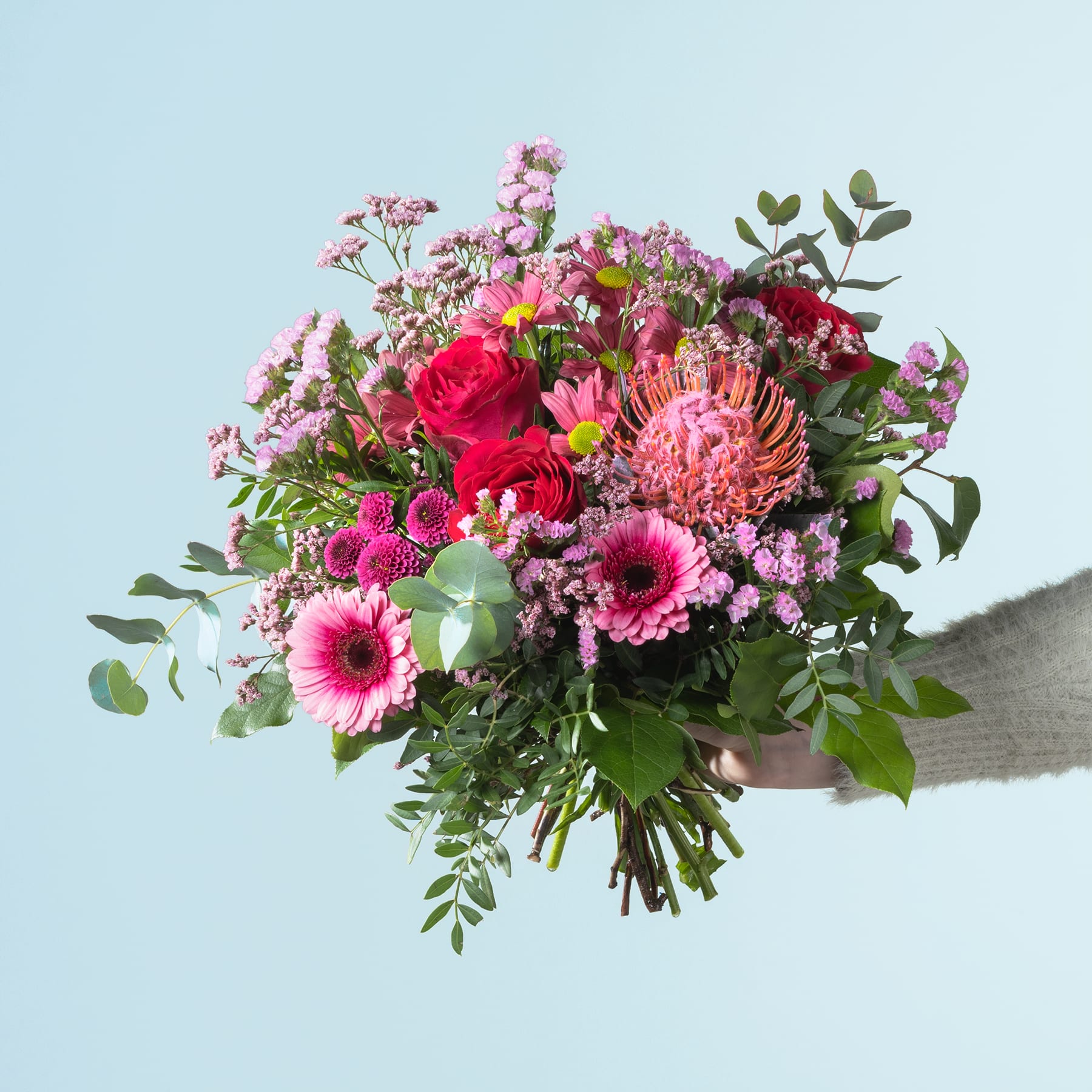 Ein farbenfroher Strauß verschiedener rosa, roter und violetter Blumen, darunter Rosen, Gerbera und Chrysanthemen, wird vor einem schlichten hellblauen Hintergrund gehalten.