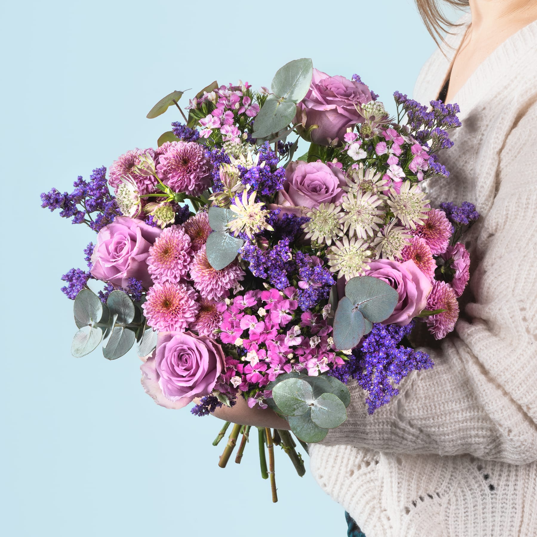 Ein leuchtender Strauß rosa und violetter Blumen, darunter Rosen, Chrysanthemen und Lavendel, wird von einer Person in den Händen gehalten, die einen grauen Pullover vor einem hellblauen Hintergrund trägt.