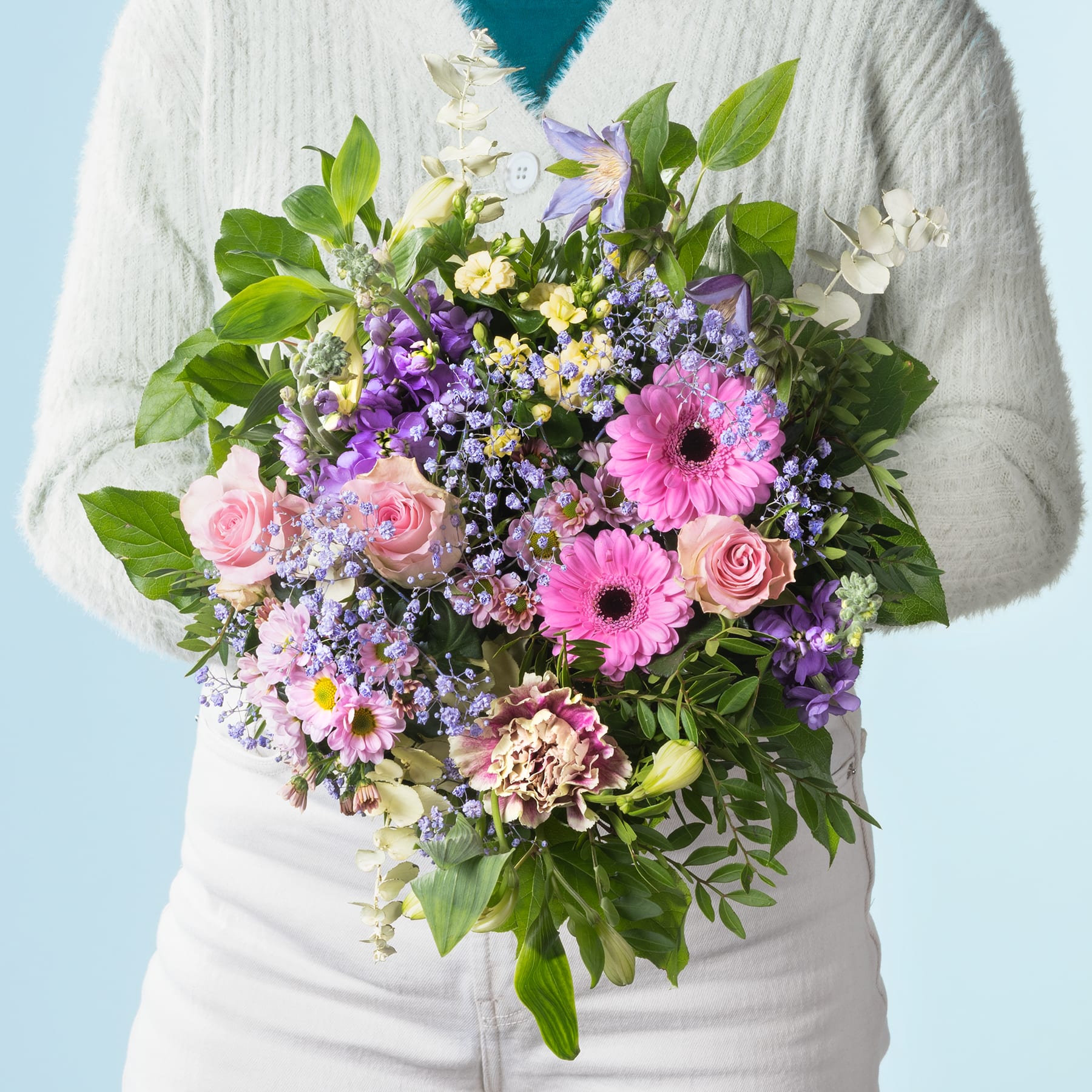 Ein lebendiger Strauß bunter Blumen, darunter rosa Gerbera, gelbe Rosen und violette und weiße Blüten, steht im Vordergrund vor einem schlichten weißen Hintergrund.