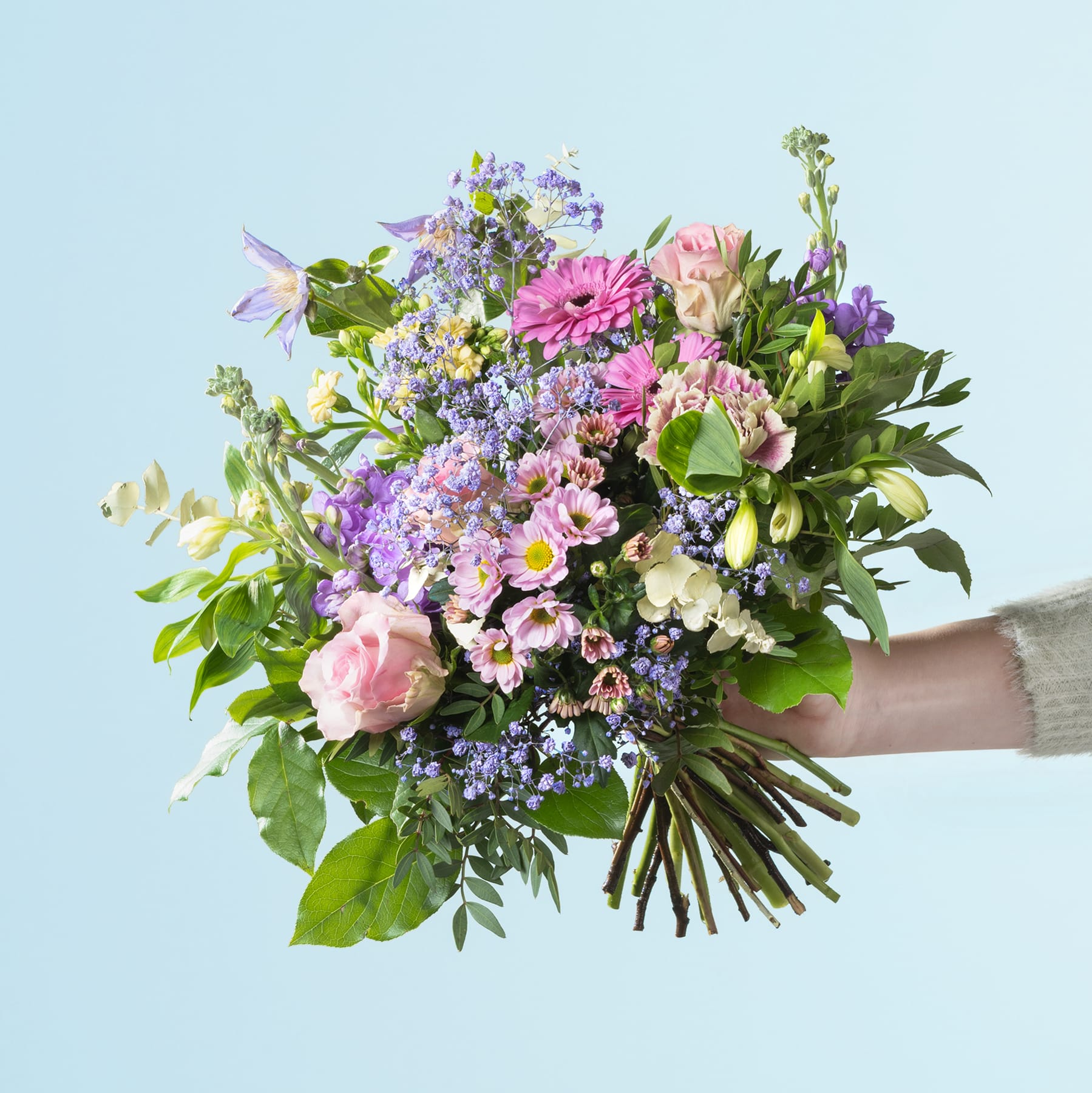 Ein lebendiger Strauß verschiedener bunter Blumen, darunter rosa Rosen, lila Flieder und grünes Laub, wird vor einem hellblauen Hintergrund in den Händen einer Person gehalten.