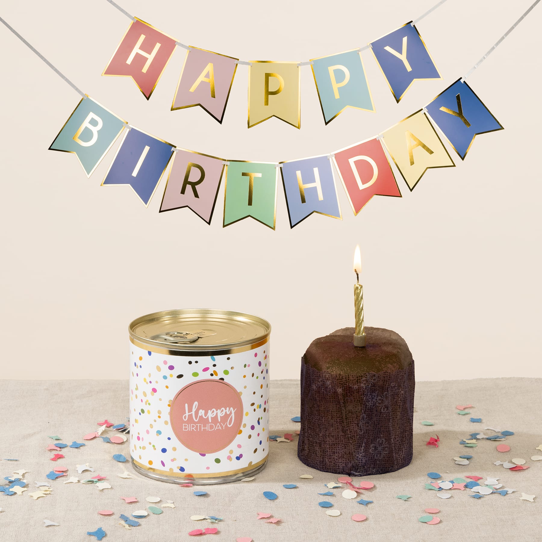Ein buntes „Happy Birthday“-Banner hängt über einer Geburtstagstorte mit einer brennenden Kerze und einer dekorierten Blechdose auf einem Tisch, auf dem Konfetti verstreut ist.