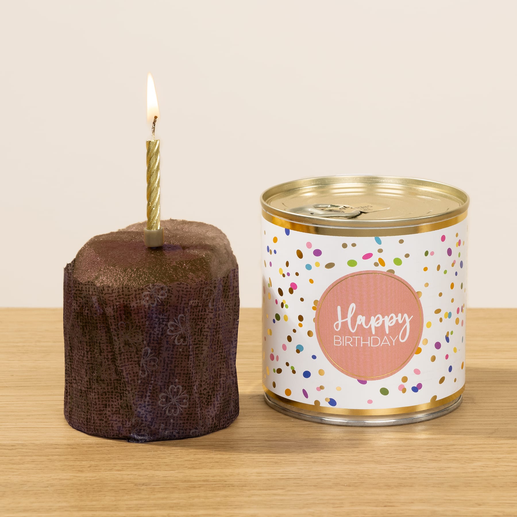 Eine Schokoladengeburtstagstorte mit einer brennenden Kerze steht neben einer bunten Blechdose mit der Aufschrift „Happy Birthday“, alles auf einer Holzoberfläche platziert.