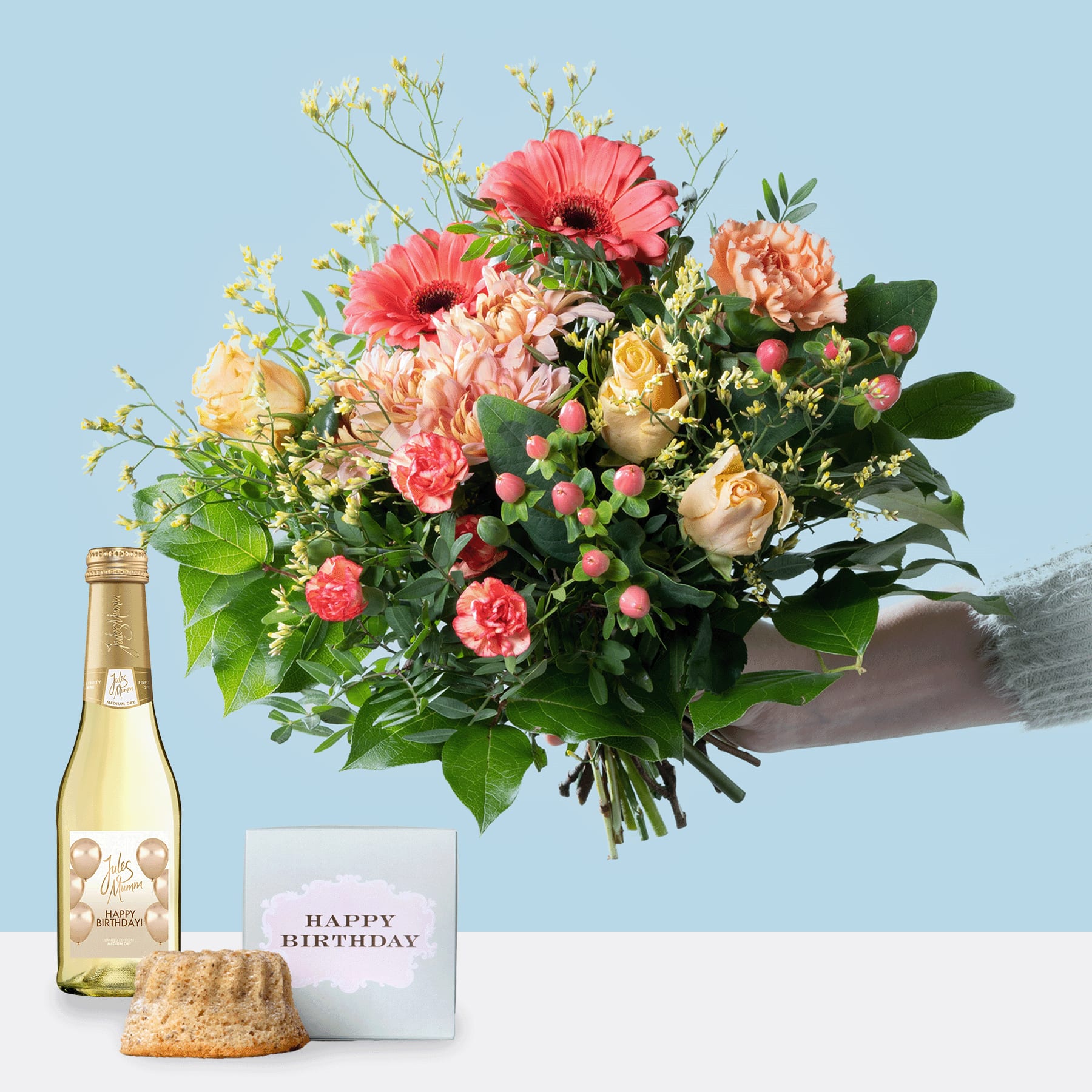 Ein leuchtender Strauß bunter Blumen, darunter rote Gerbera, pfirsichfarbene Rosen und Grünpflanzen, wird vor einer Flasche Weißwein und einer Geburtstagskarte auf einer weißen Fläche vor einem hellblauen Hintergrund gehalten.