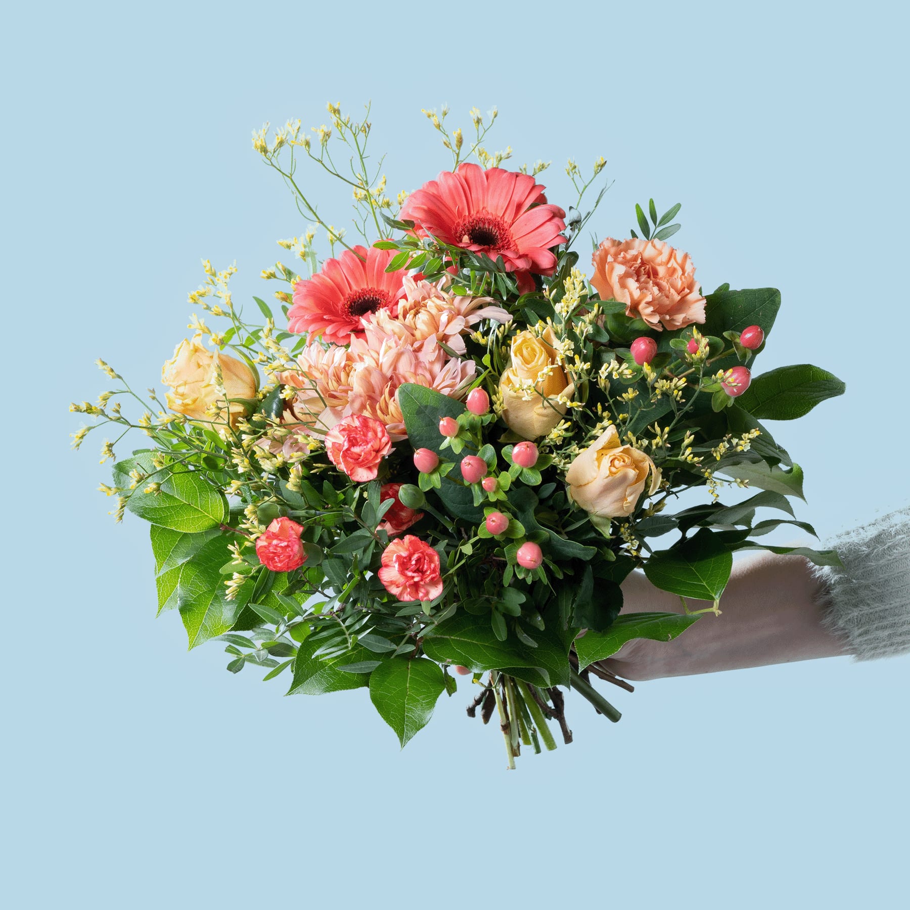 Ein lebendiger Strauß bunter Blumen, darunter rosa Gerbera, pfirsichfarbene Rosen und Grünpflanzen, wird vor einem schlichten blauen Hintergrund in den Händen einer Person gehalten.