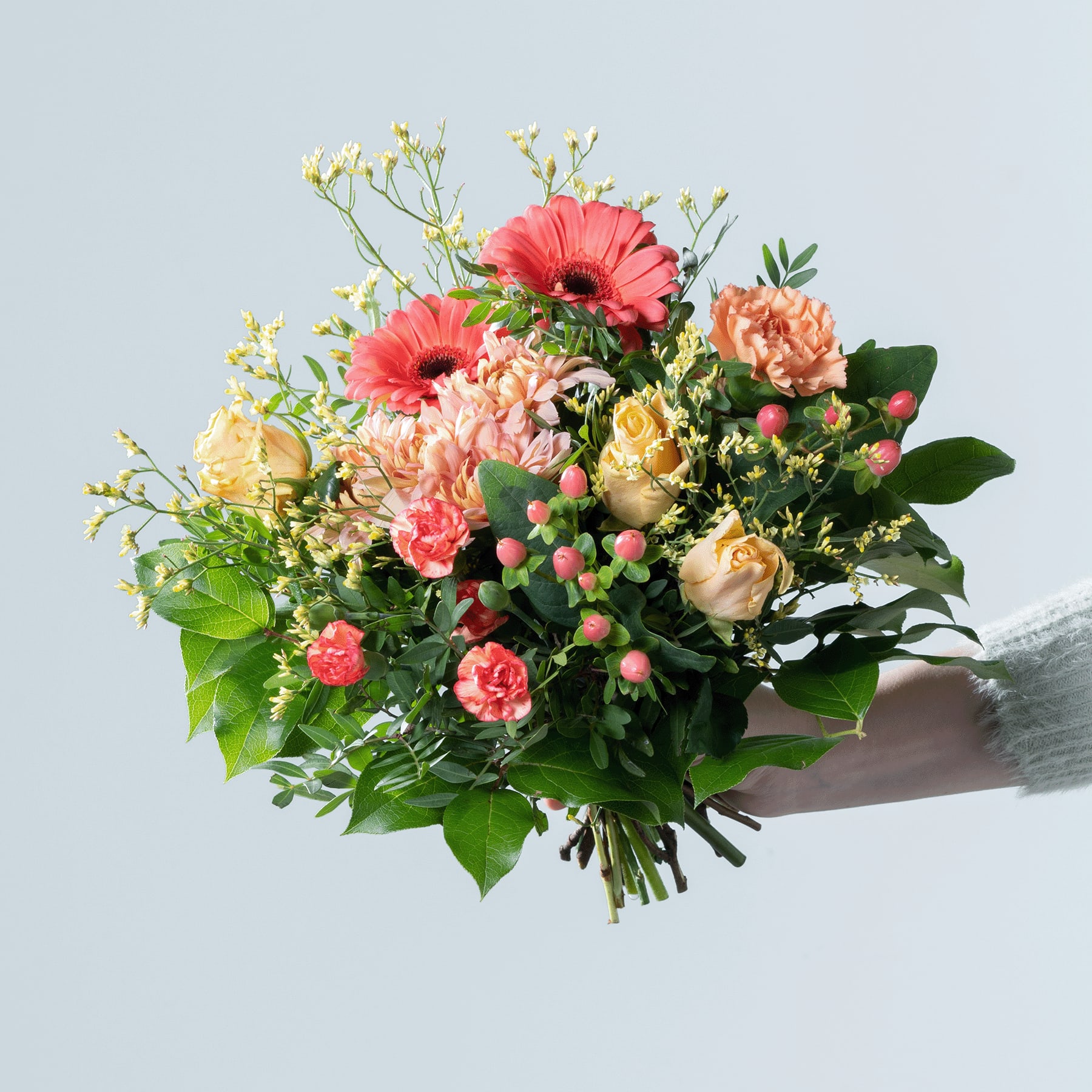 Ein lebendiger Strauß bunter Blumen, darunter rosa Gerbera, gelbe Rosen und verschiedene Grünpflanzen, wird vor einem schlichten Hintergrund in den Händen einer Person gehalten.