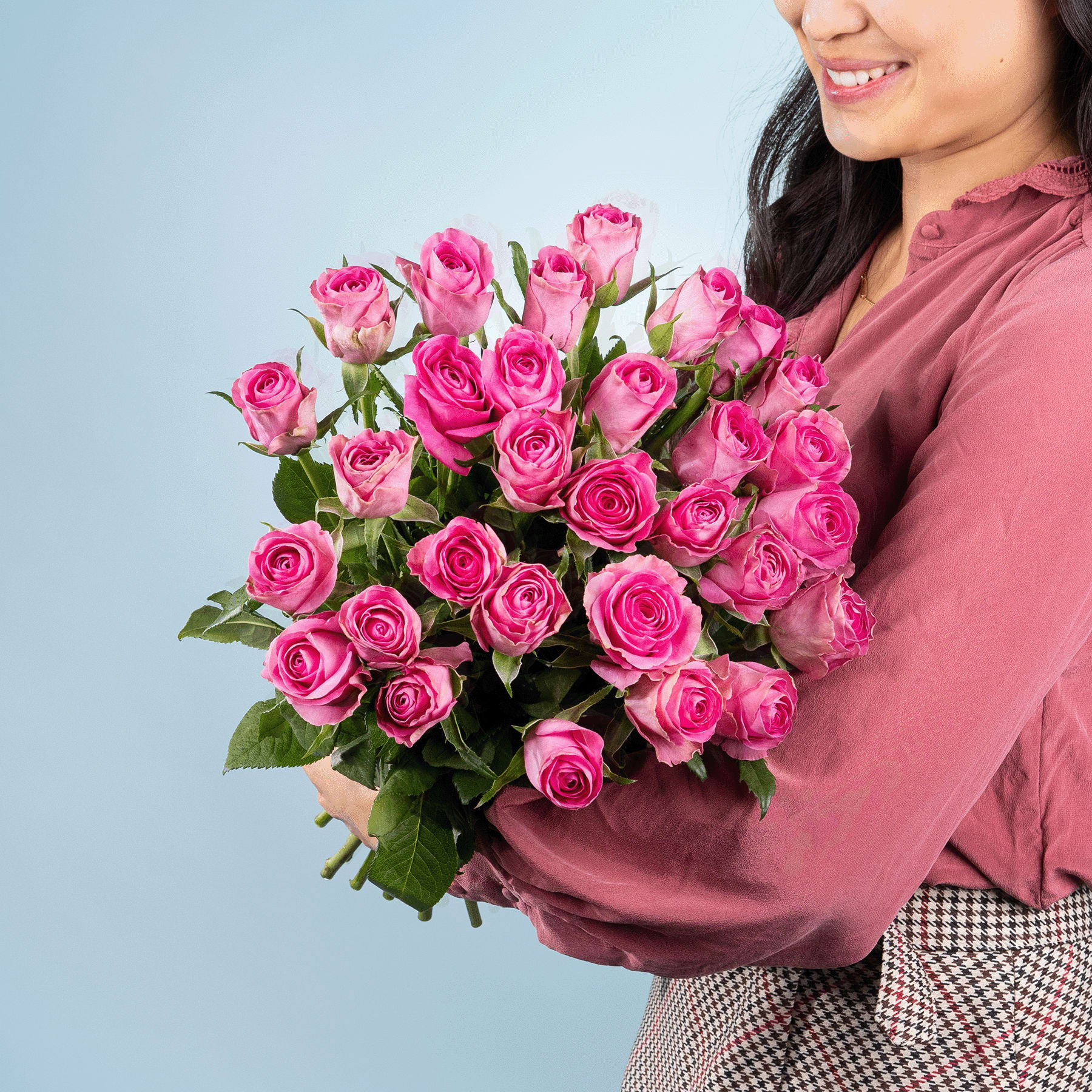 Eine Frau in einem rosa Hemd hält einen großen Strauß leuchtend rosa Rosen vor einem schlichten blauen Hintergrund.