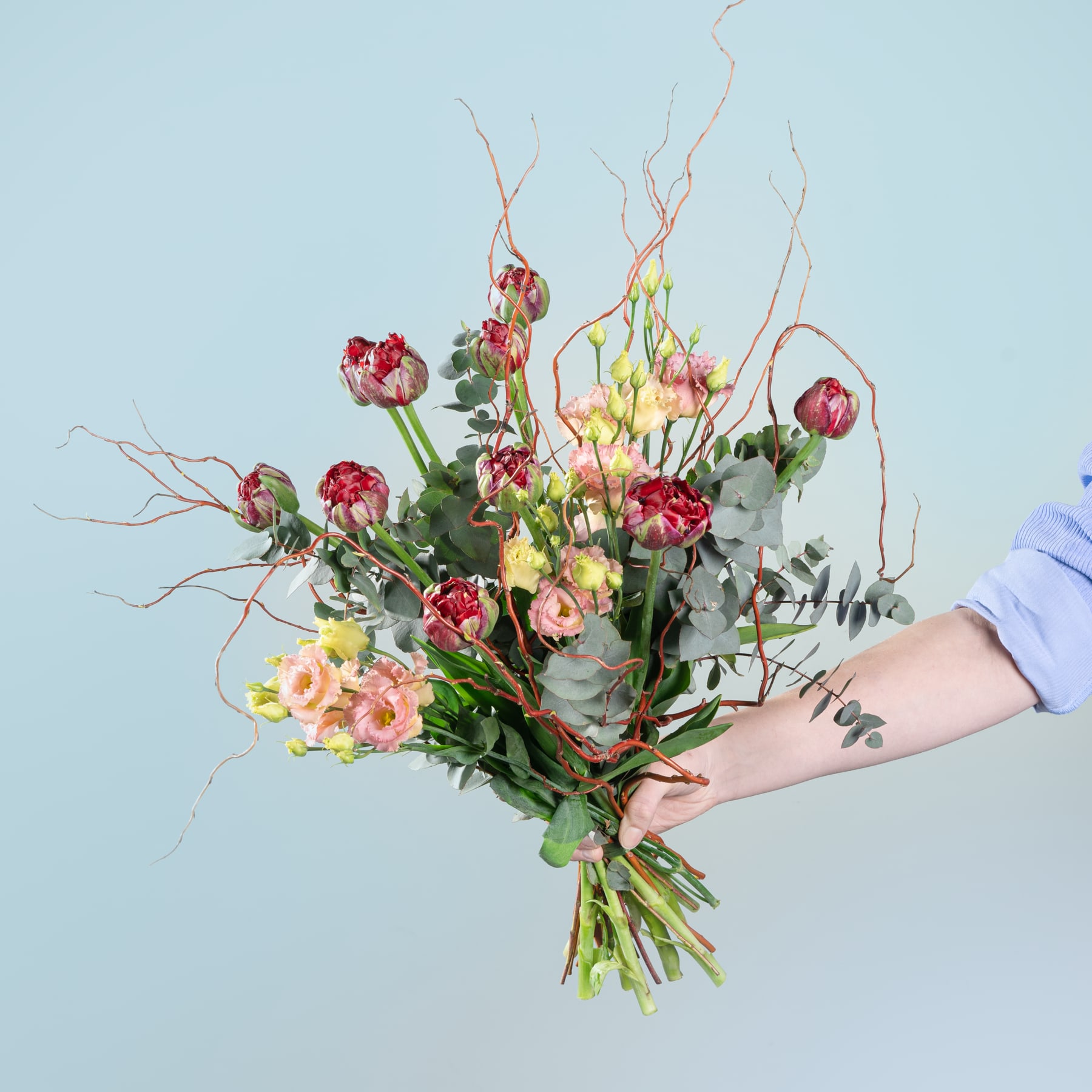 Ein farbenfroher Strauß verschiedener Blumen, darunter Rosen, Tulpen und andere Blüten, den eine Person vor einem schlichten blauen Hintergrund in den Händen hält.