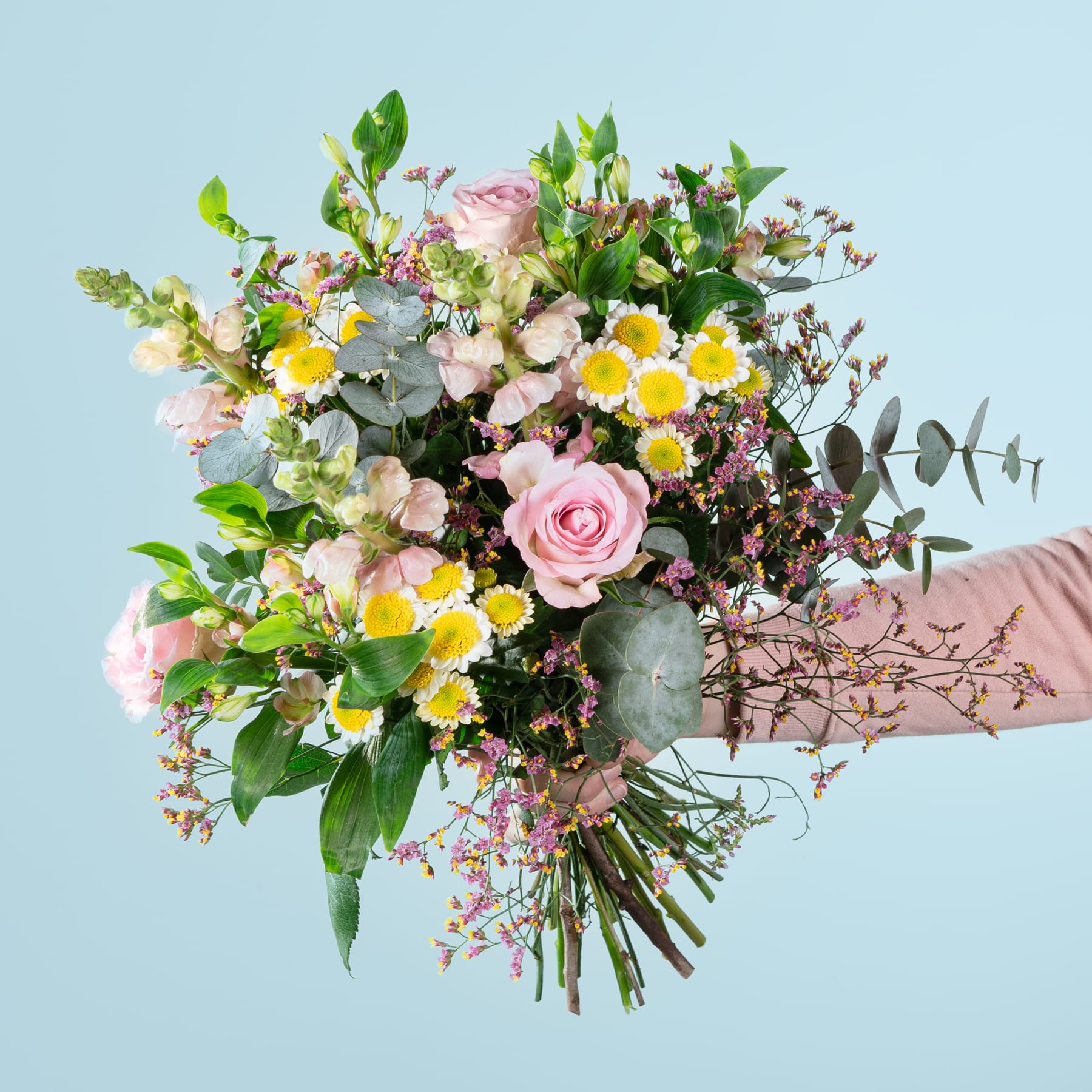 Ein farbenfroher Strauß verschiedener Blumen, darunter rosa Rosen, gelbe Gänseblümchen und Grünpflanzen, wird vor einem hellblauen Hintergrund in den Händen einer Person gehalten.