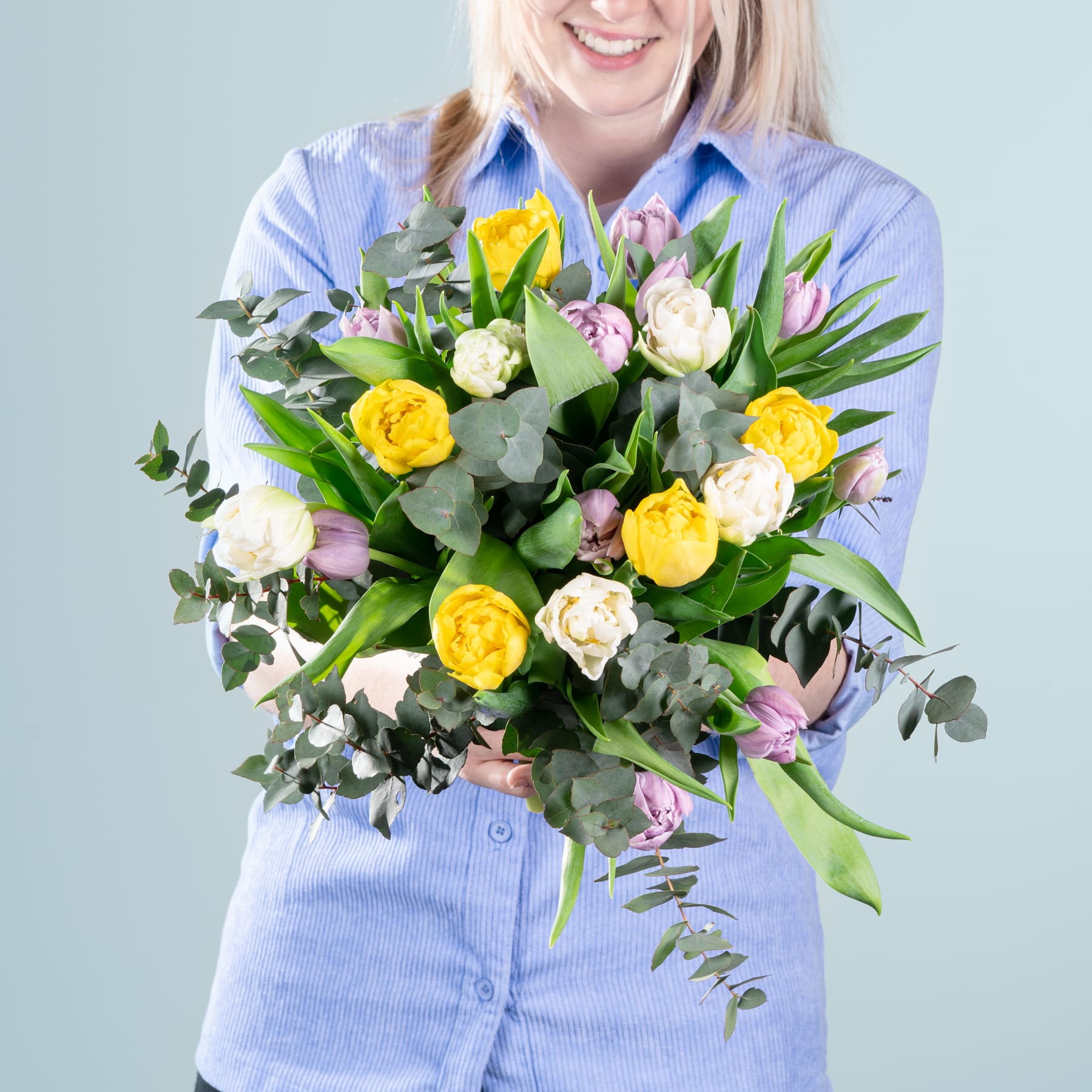 Eine lächelnde Frau in einem blauen Hemd hält einen farbenfrohen Strauß gelber und weißer Blumen vor einem schlichten hellblauen Hintergrund.