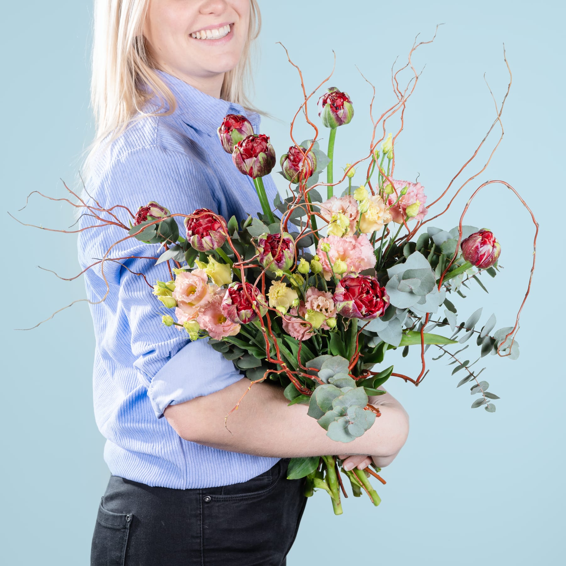 Eine lächelnde Frau in einem blauen Pullover hält vor einem schlichten blauen Hintergrund einen leuchtend bunten Blumenstrauß, darunter Rosen, Narzissen und Grünpflanzen.