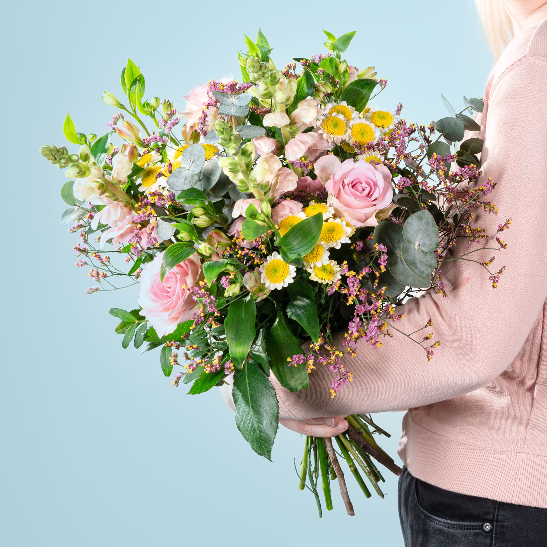 Ein farbenfroher Strauß verschiedener Blumen, darunter rosa Rosen, Gänseblümchen und Grünpflanzen, wird im Vordergrund vor einem sanften blauen Hintergrund gehalten.