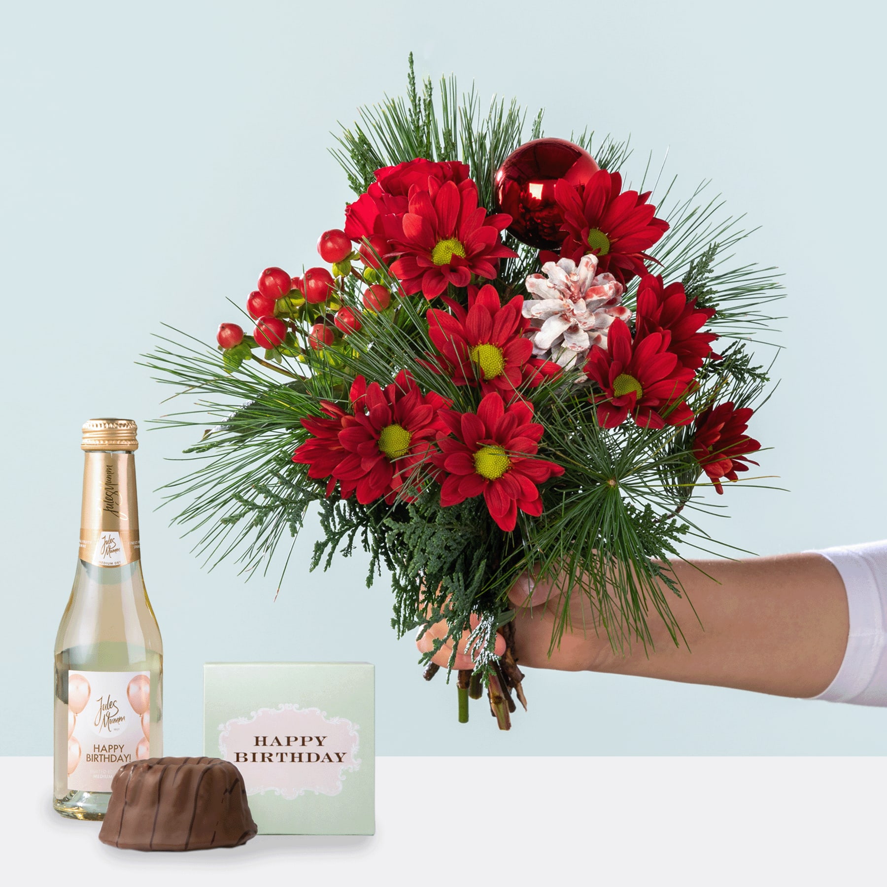 Ein farbenfroher Strauß roter und rosa Blumen, darunter Chrysanthemen und Tannenzweige, wird vor einem schlichten Hintergrund in einer Hand gehalten, begleitet von einer Flasche Wein und einer Geburtstagskarte.