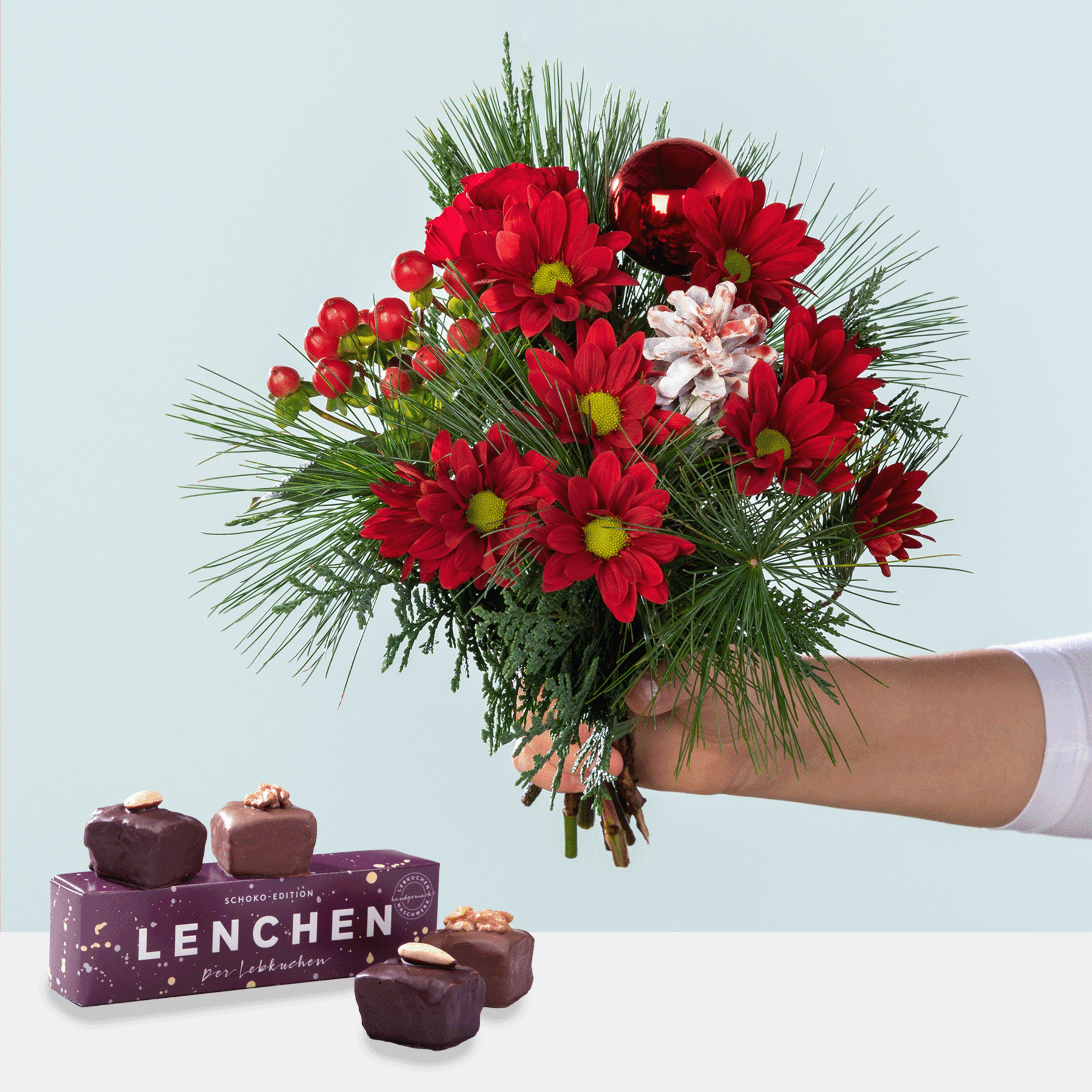 Ein farbenfroher Strauß roter und rosa Blumen, darunter Gänseblümchen und andere Blüten, wird vor einem schlichten Hintergrund in einer Hand gehalten, begleitet von einer Schachtel Pralinen mit der Aufschrift „Lenchen“.