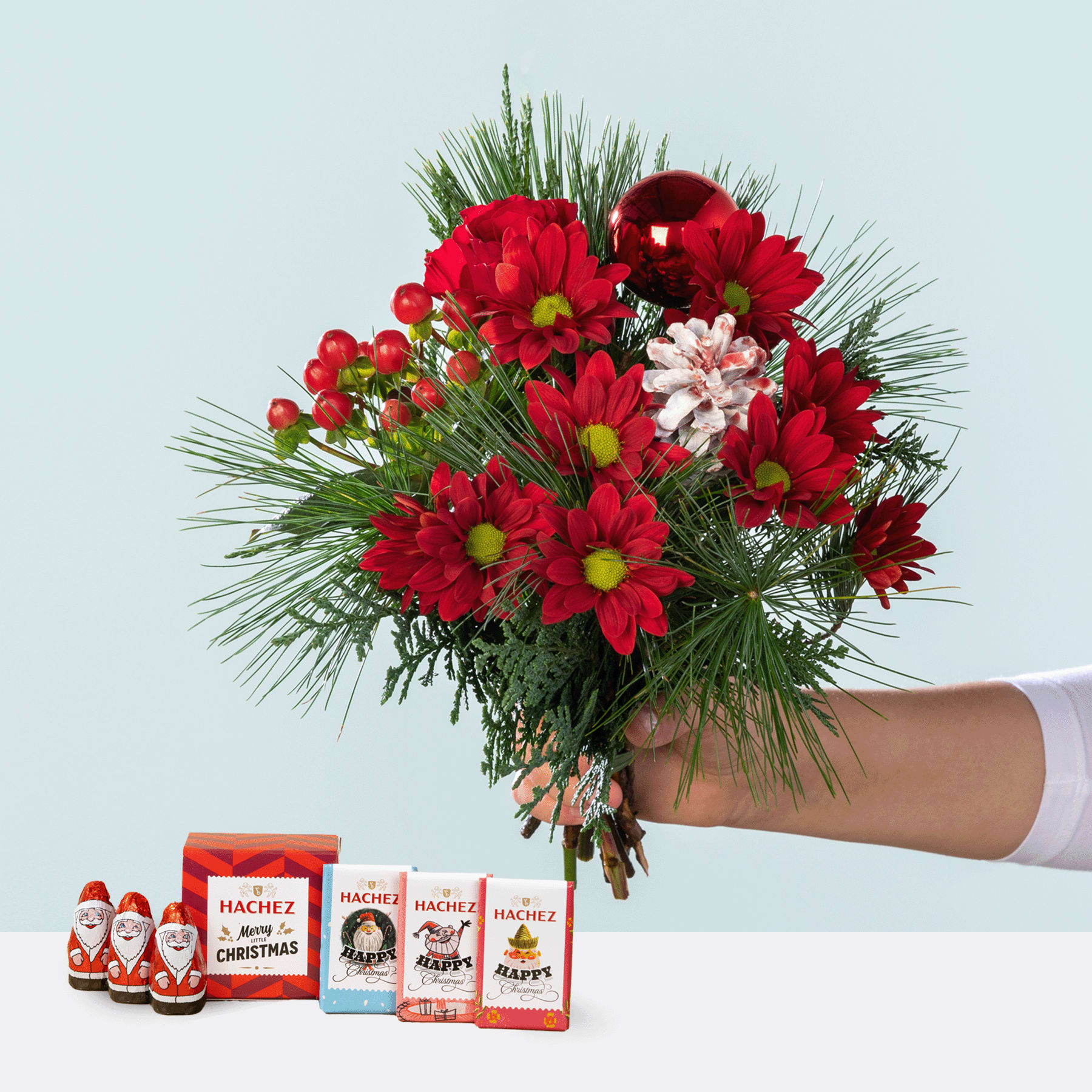 Ein leuchtender Strauß roter und rosa Blumen, darunter Chrysanthemen und andere Blüten, wird vor einem schlichten Hintergrund in einer Hand gehalten. In der Nähe sind mehrere kleine Weihnachtsmannfiguren zu sehen.