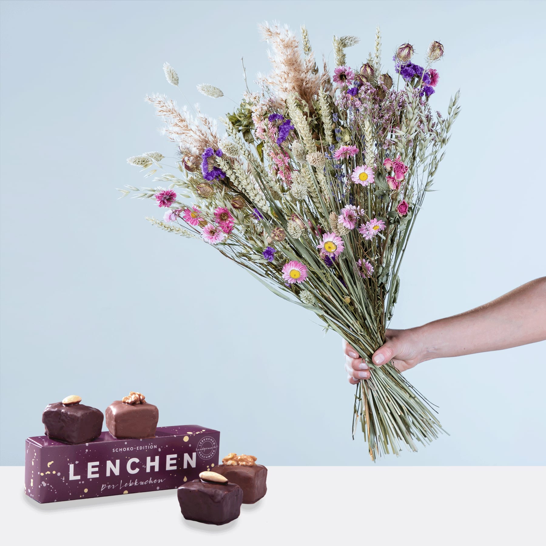 In einer Hand hält er einen Strauß leuchtend violetter und rosa Wildblumen, dazu eine Schachtel Pralinen mit der Aufschrift „Lenchen“ auf weißer Fläche vor hellblauem Hintergrund.