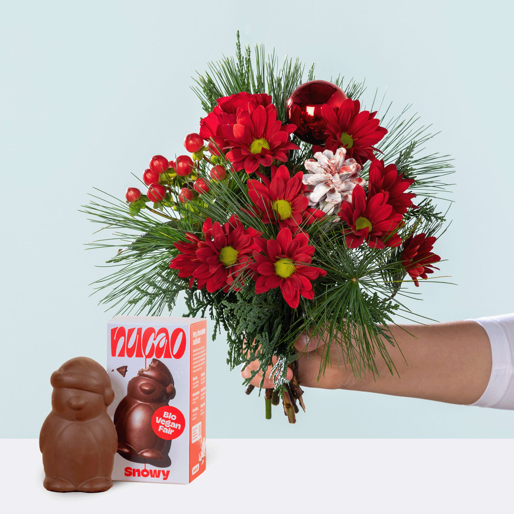 Ein leuchtender Strauß roter und rosa Blumen, darunter Chrysanthemen und Nelken, wird vor einem schlichten Hintergrund in einer Hand gehalten. Im Vordergrund ist eine Schachtel mit schokoladenüberzogenen Marshmallow-Leckereien zu sehen.