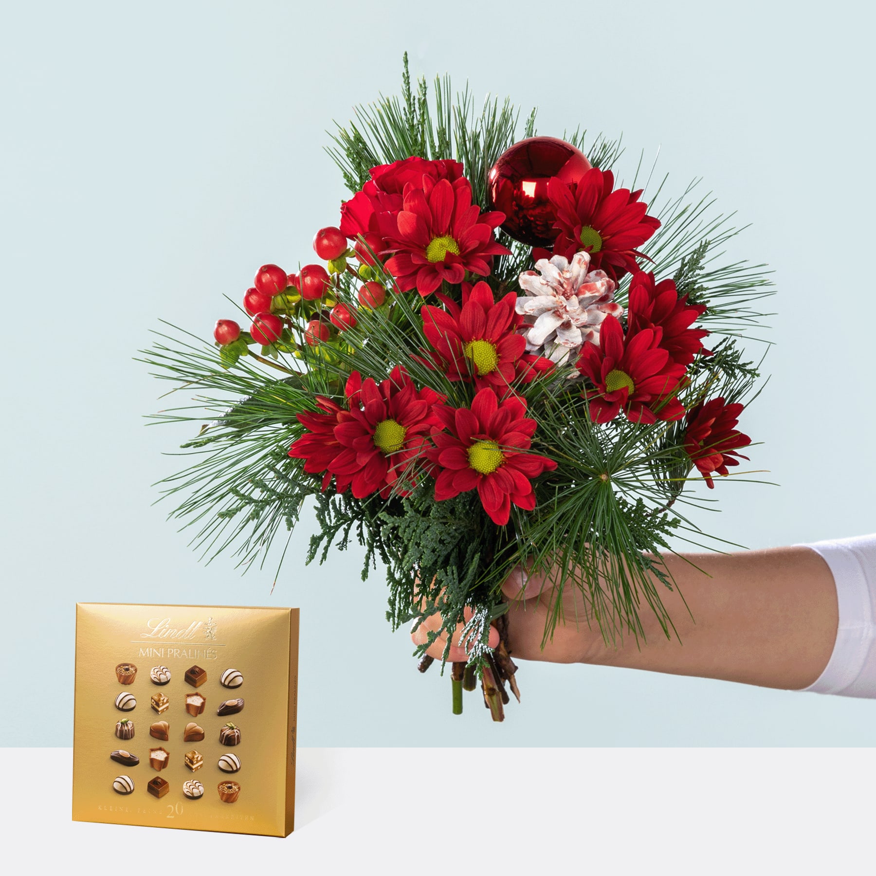Ein leuchtender Strauß roter und rosa Blumen, darunter Chrysanthemen und Tannenzweige, wird vor einem schlichten Hintergrund in einer Hand gehalten, neben einer goldenen Schachtel Pralinen.