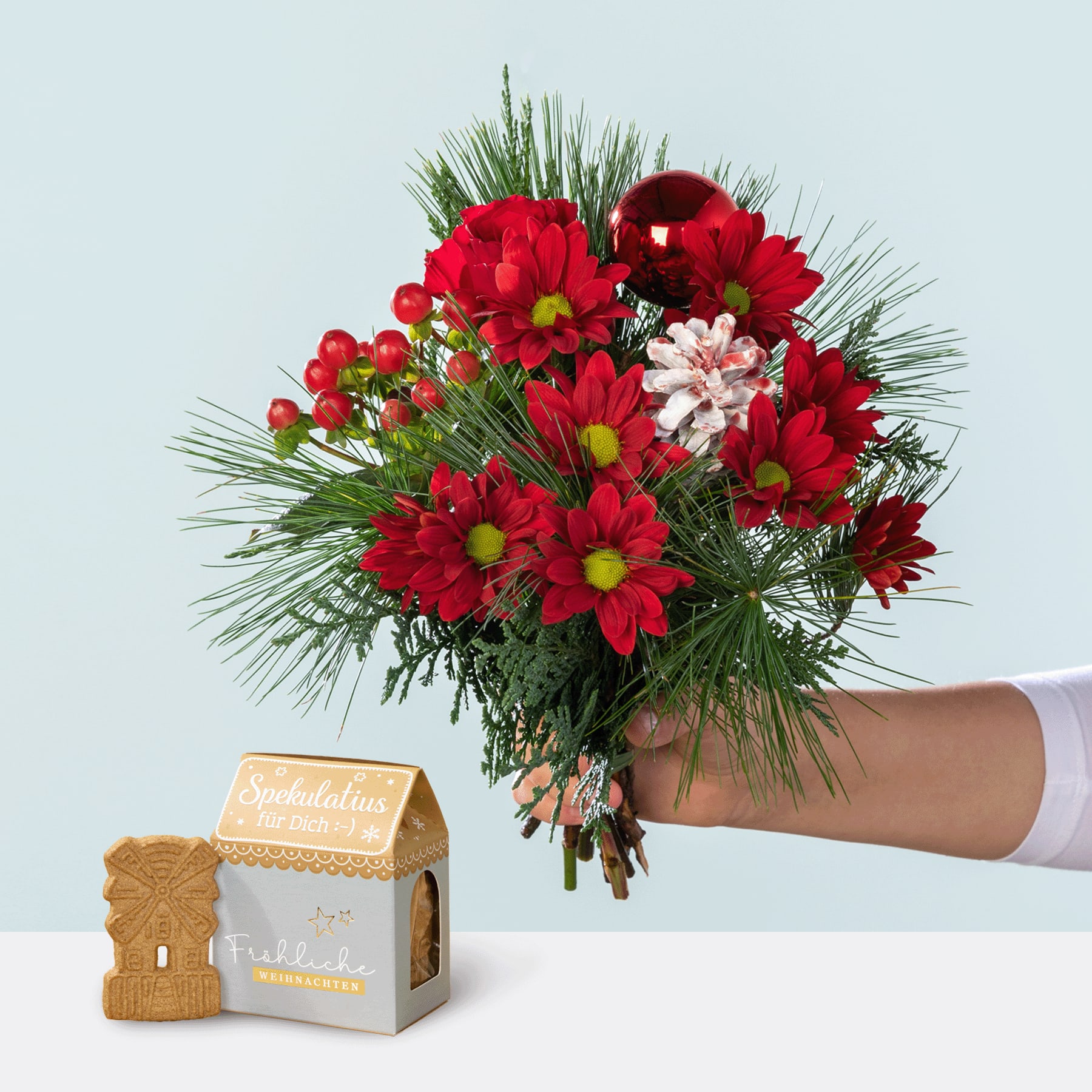 Ein farbenfroher Strauß roter und rosa Blumen, darunter Chrysanthemen und andere Blüten, wird vor einem schlichten Hintergrund in einer Hand gehalten, begleitet von einem kleinen hölzernen Objekt in Form eines Hauses.