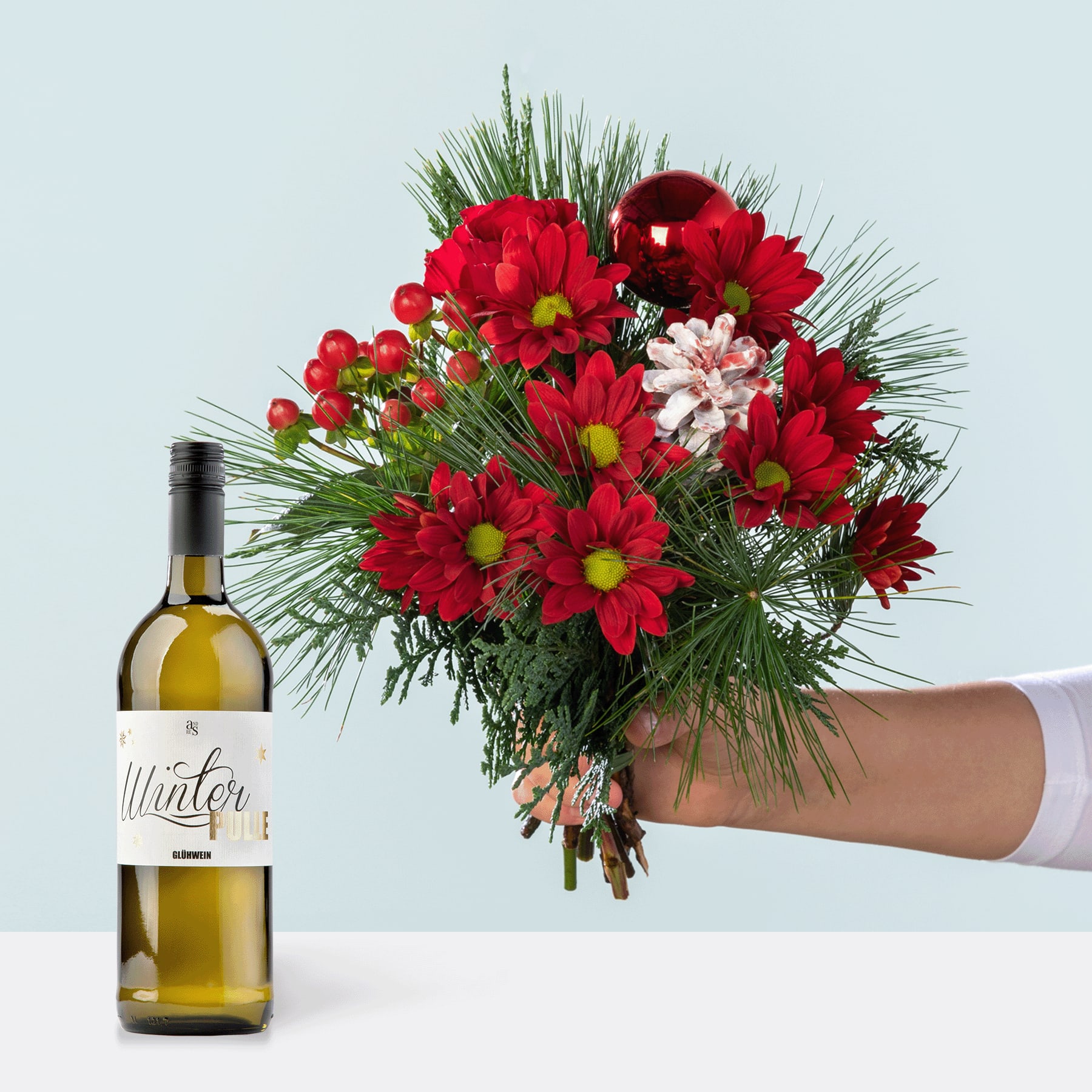 Ein farbenfroher Strauß roter und rosa Blumen, darunter Chrysanthemen und Nelken, wird neben einer Flasche Weißwein vor einem schlichten Hintergrund in einer Hand gehalten.