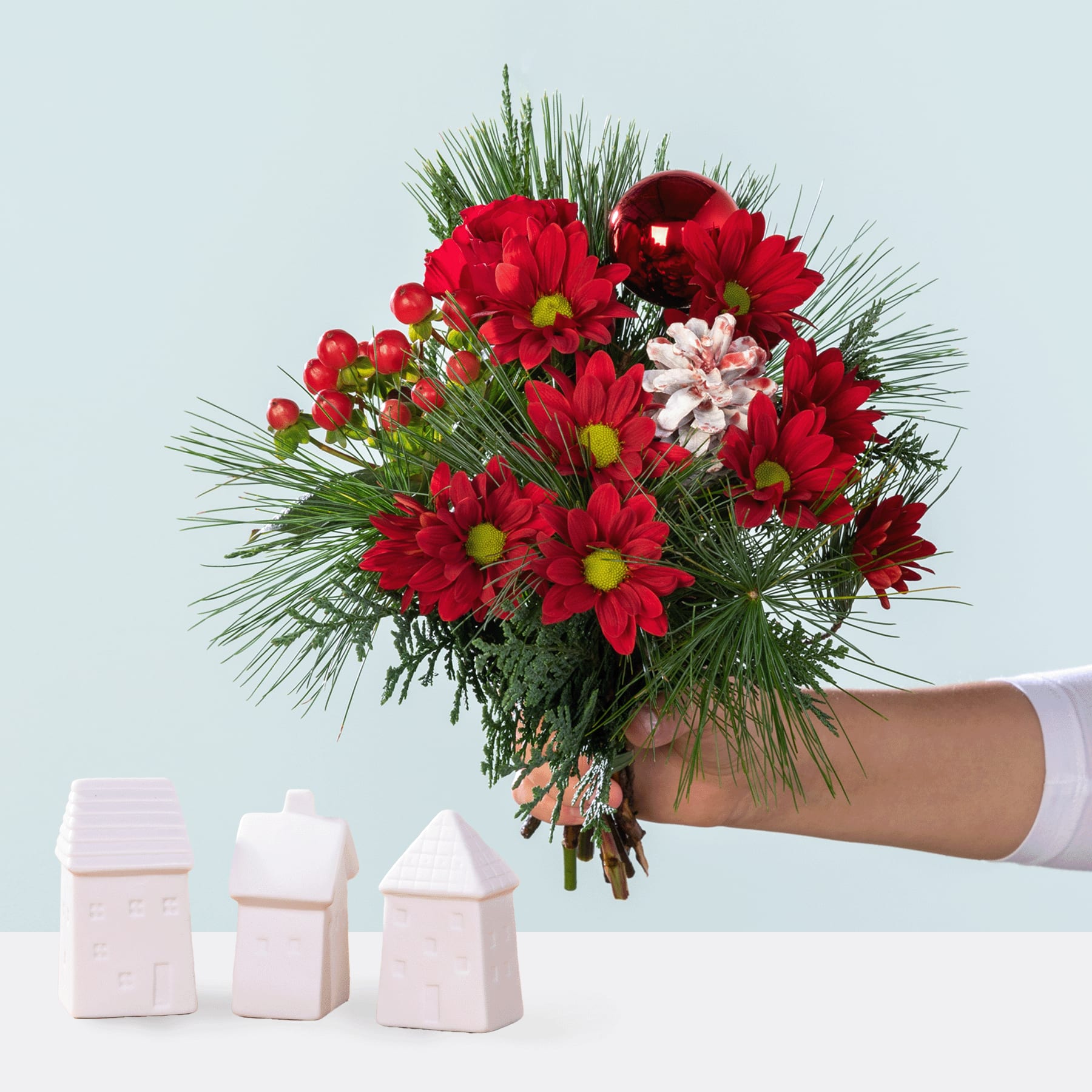 Ein leuchtender Strauß roter und rosa Blumen, darunter Chrysanthemen und andere Blüten, in einer Hand gehalten vor einem schlichten Hintergrund mit kleinen weißen Dekorationsobjekten.