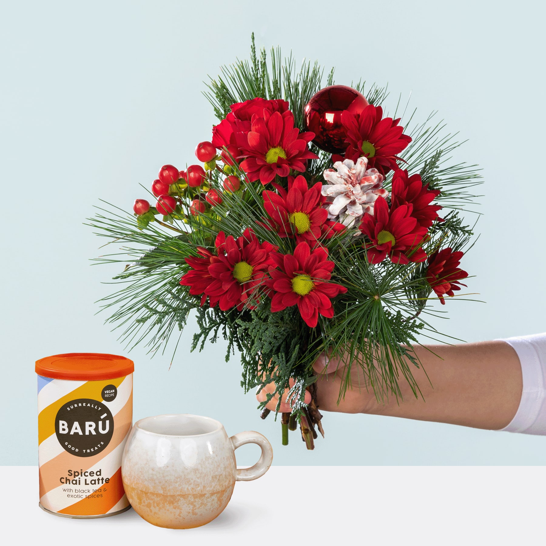 Ein farbenfroher Strauß roter und rosa Blumen, darunter Chrysanthemen und Nelken, wird vor einem schlichten Hintergrund in einer Hand gehalten, begleitet von einer Dose Chai Latte mit Baru-Gewürzen.