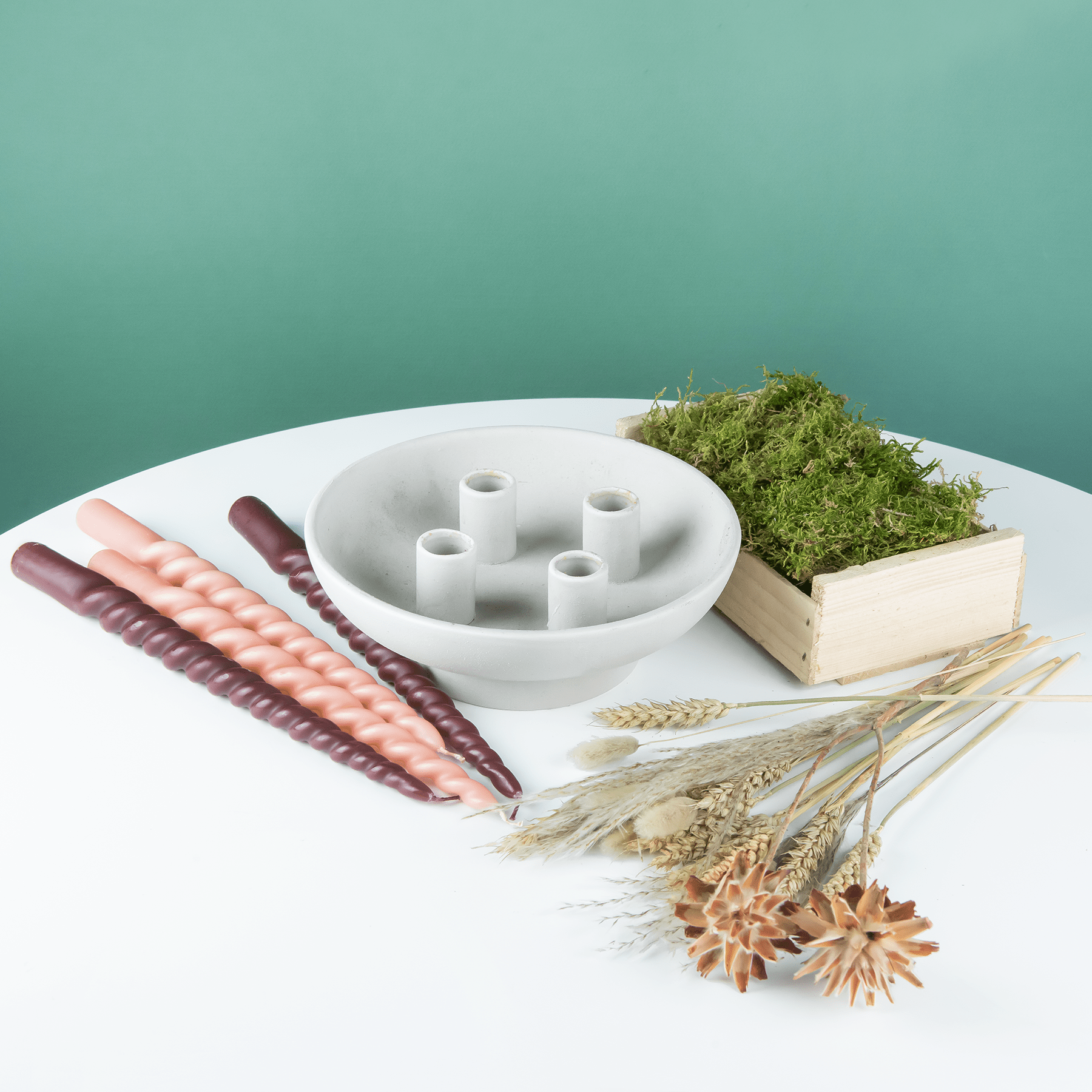 Ein weißer Keramikteller mit verschiedenen Trockenblumen, eine mit Moos gefüllte Holzkiste und ein Satz Holzstäbe auf einer schlichten weißen Fläche vor einem mintgrünen Hintergrund.