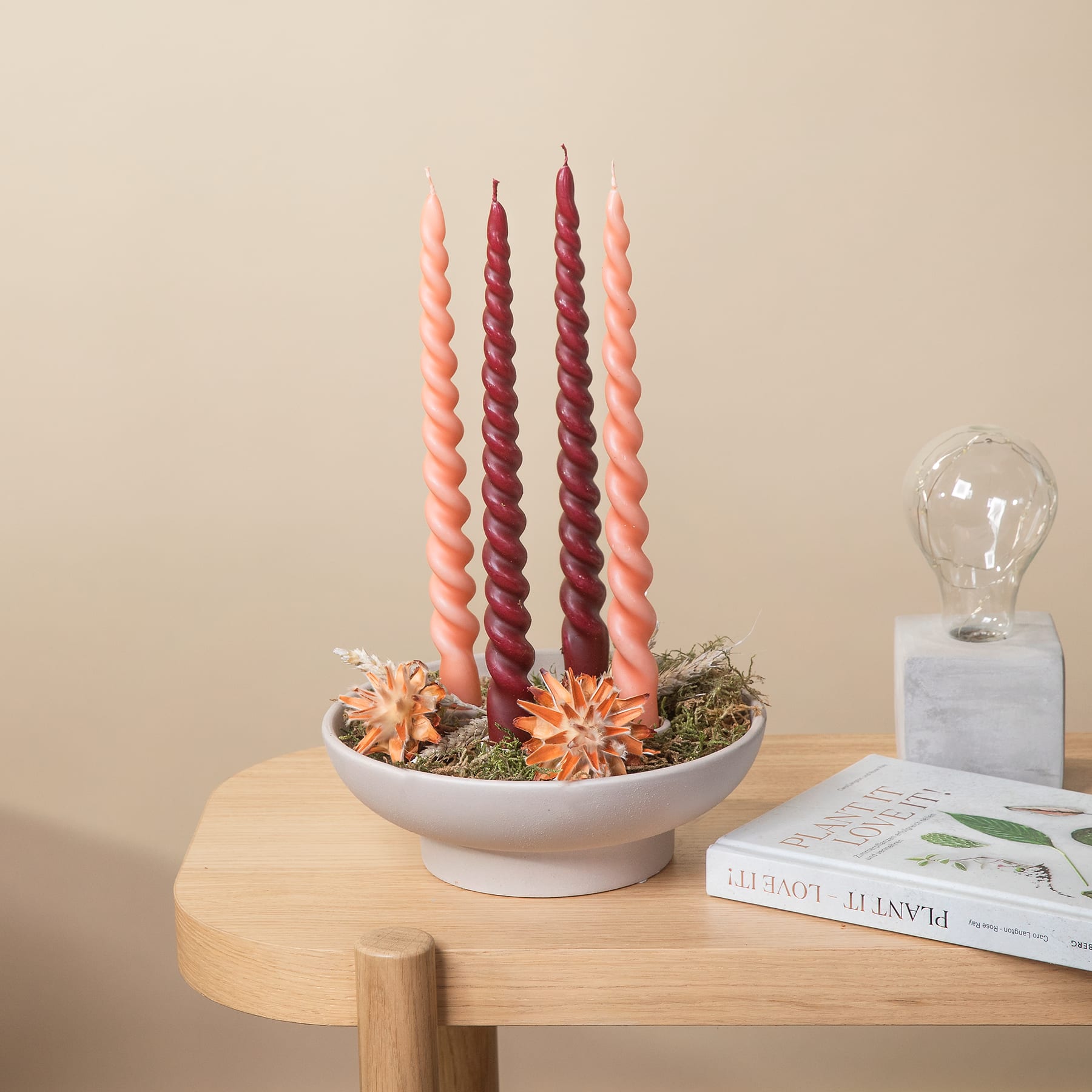 Auf einem Holztisch steht eine Schale mit bunten Kerzen und Sukkulenten, im Hintergrund eine Glaslampe und Bücher.