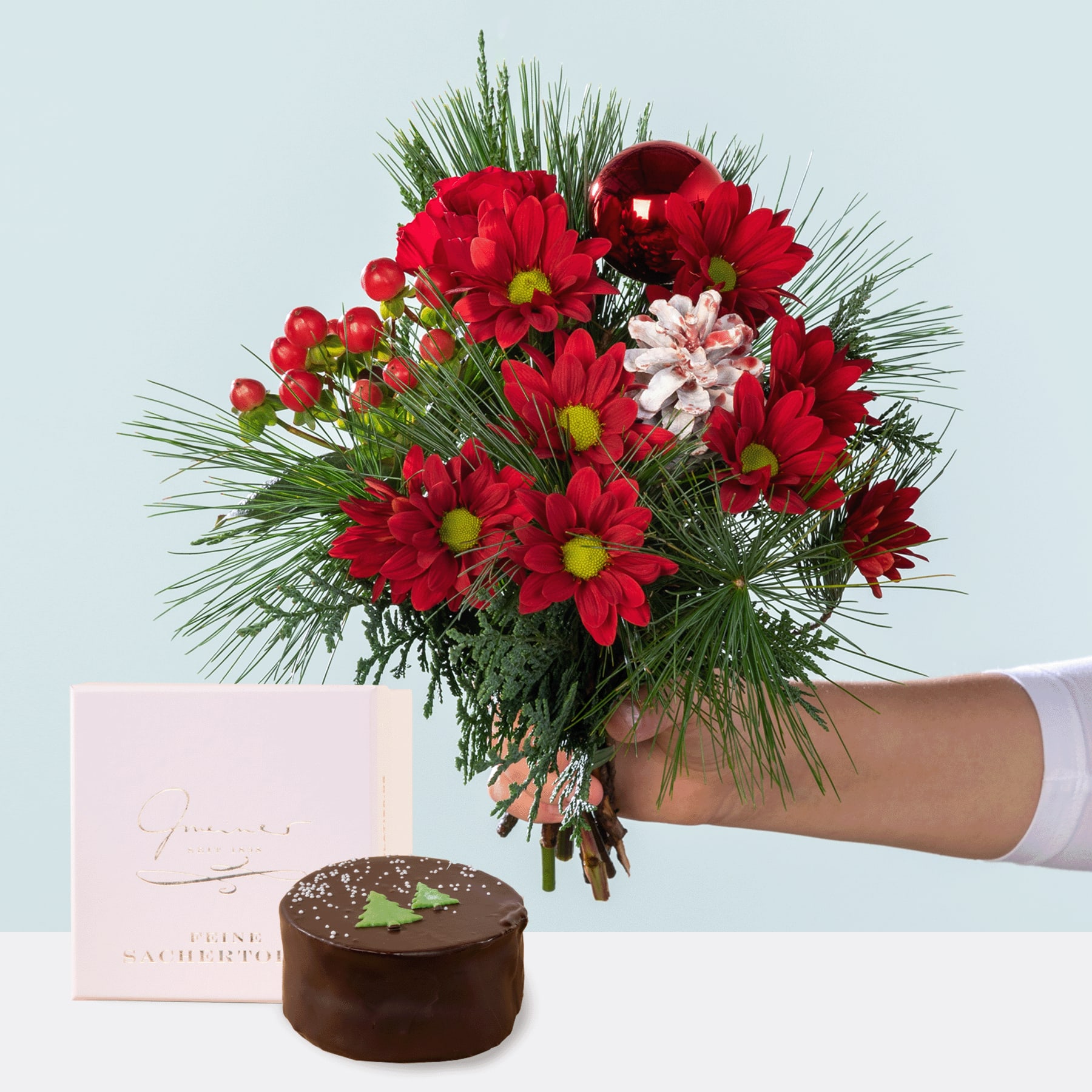 Ein farbenfroher Strauß roter und weißer Blumen, darunter Gänseblümchen und Tannenzweige, wird vor einem schlichten Hintergrund in einer Hand gehalten, begleitet von einem kleinen Schokoladenkuchen.
