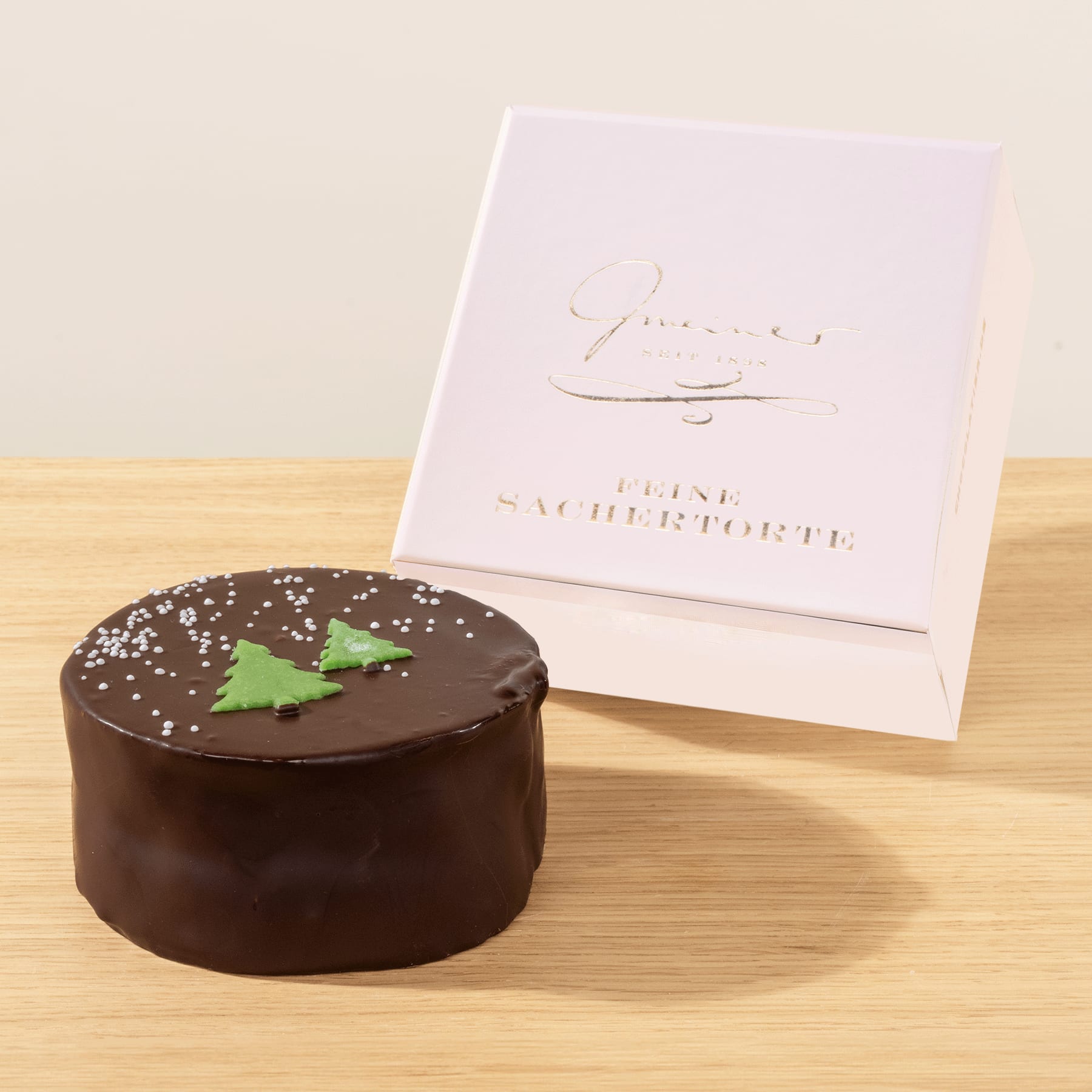 Auf einer Holzoberfläche steht ein Schokoladenkuchen mit einer grünen baumförmigen Dekoration, begleitet von einer weißen Geschenkbox mit einem Logo in Schreibschrift.