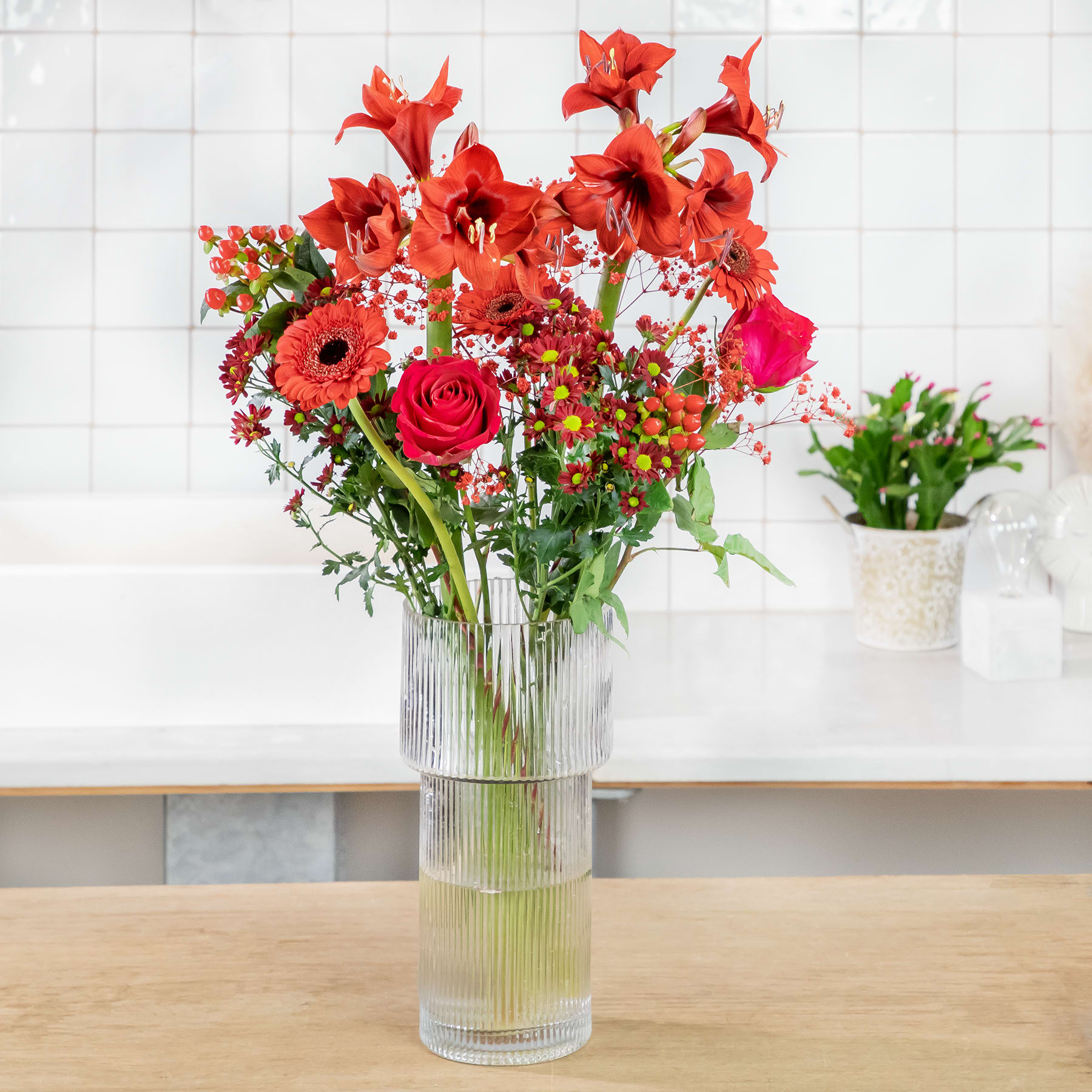 Ein farbenfroher Strauß roter Blumen, darunter Rosen, Gerbera und andere Blüten, wird in einer Glasvase auf einem Holztisch vor dem Hintergrund einer gefliesten Wand präsentiert.