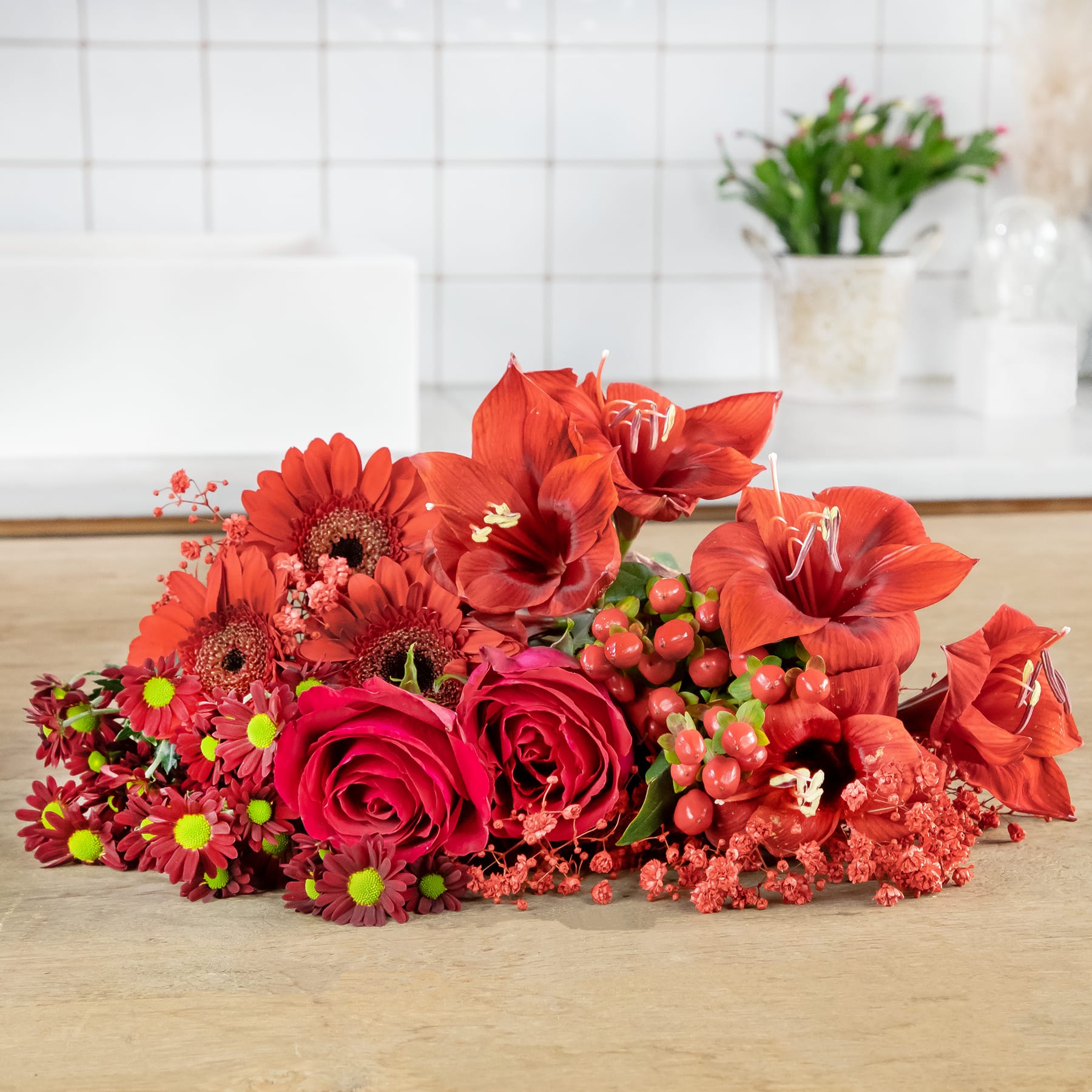 Ein farbenfroher Strauß roter Rosen, Gerbera und anderer roter Blumen steht auf einer Holzoberfläche vor dem Hintergrund einer gefliesten Wand.