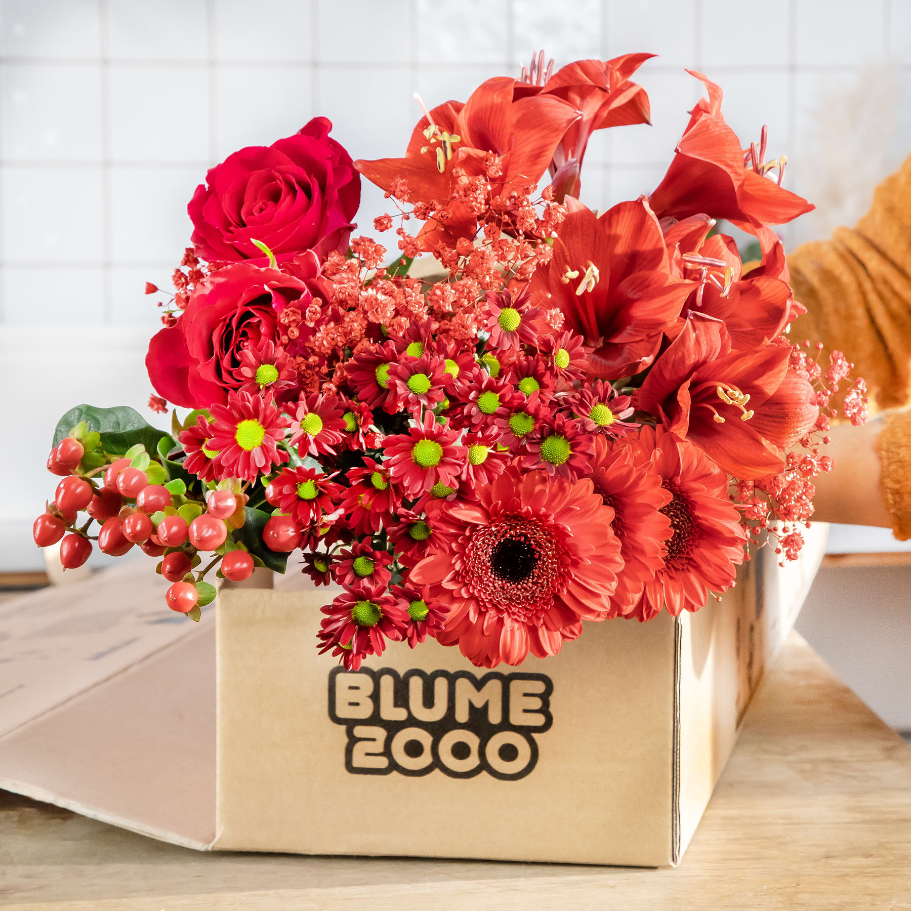 Ein farbenfroher Strauß roter Rosen, Gerbera und anderer roter Blumen in einem Karton mit der Aufschrift „Blume Coco“ steht auf einer Holzoberfläche vor einem weiß gefliesten Hintergrund.