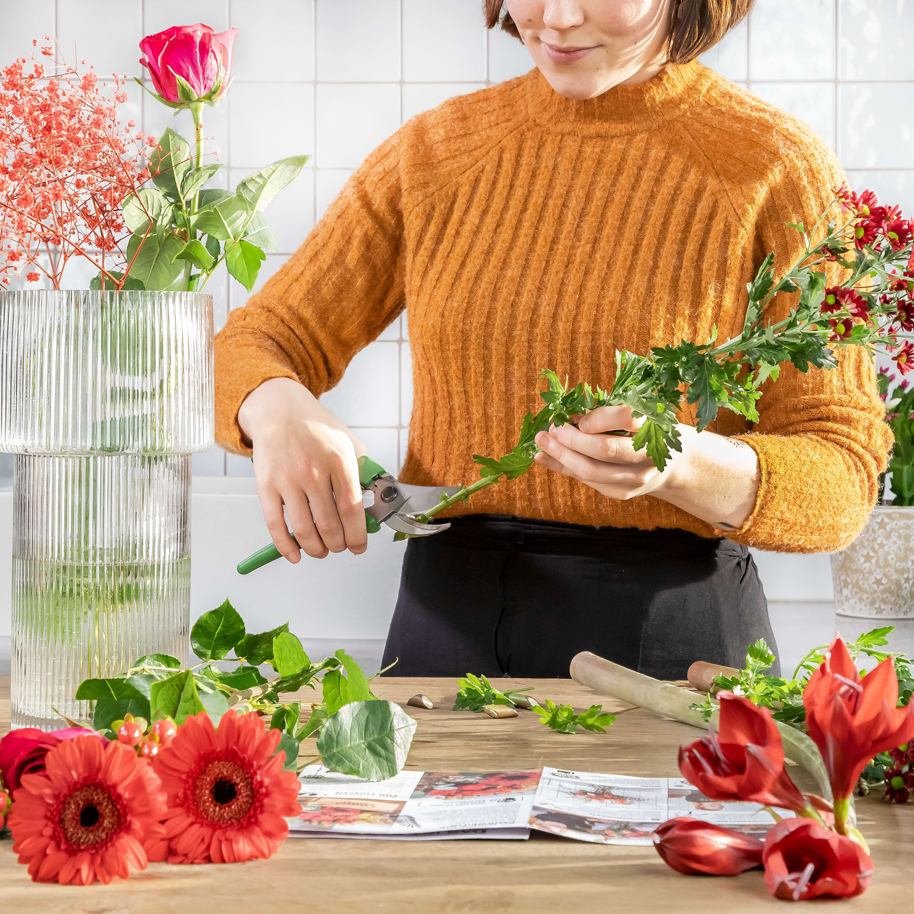 Eine Frau in einem kuscheligen senffarbenen Pullover arrangiert frische Blumen und Grünpflanzen auf einem Tisch, umgeben von leuchtend roten Gerbera-Gänseblümchen und anderen floralen Elementen.