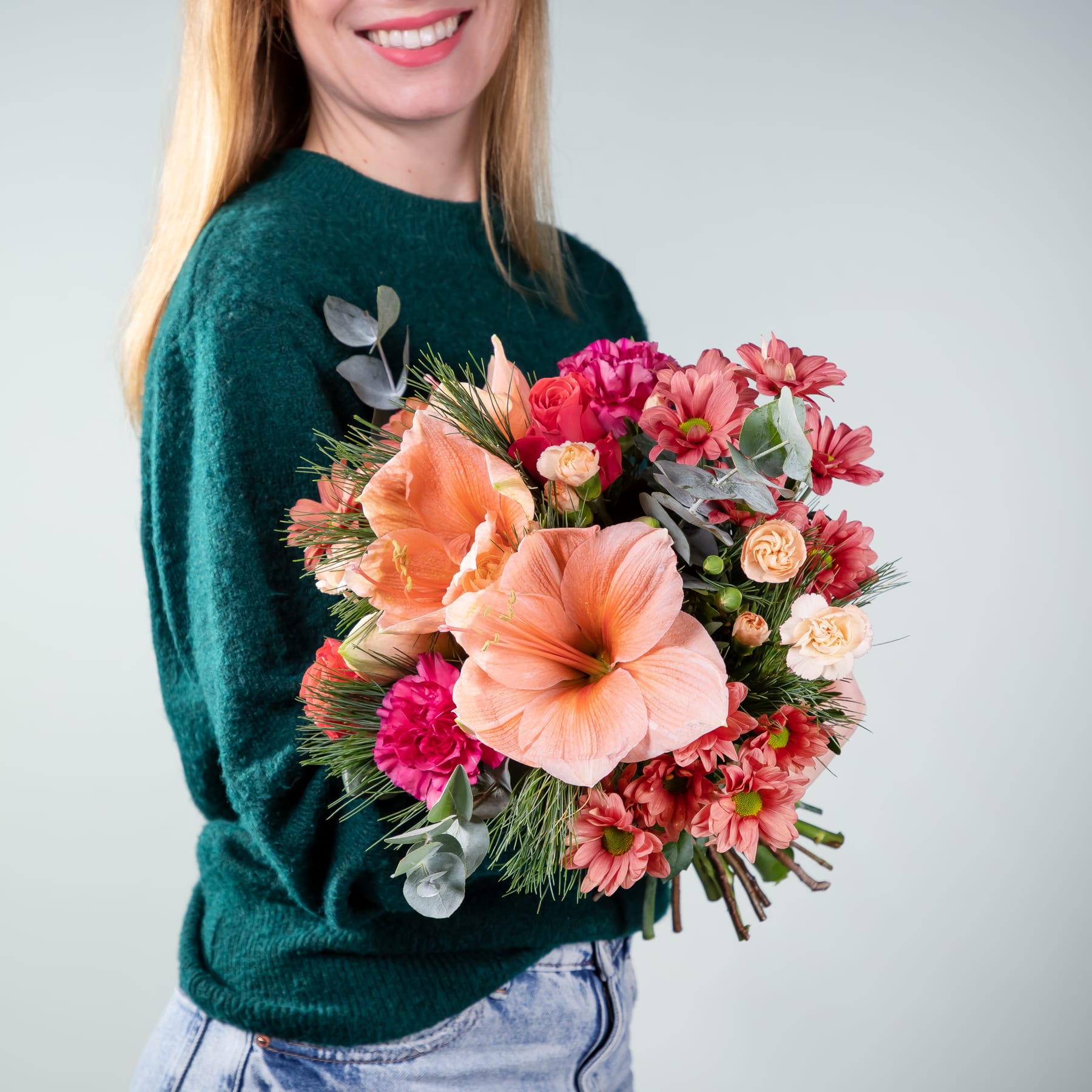 Eine lächelnde Frau in einem grünen Pullover hält einen farbenfrohen Strauß rosa und orangefarbener Blumen vor einem schlichten Hintergrund.