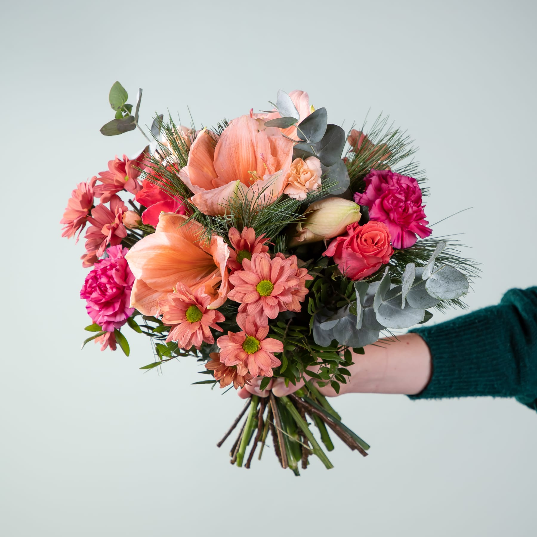 Ein farbenfroher Strauß verschiedener rosa, orangefarbener und roter Blumen, darunter Rosen, Gänseblümchen und Pfingstrosen, wird vor einem schlichten hellblauen Hintergrund in den Händen einer Person gehalten.